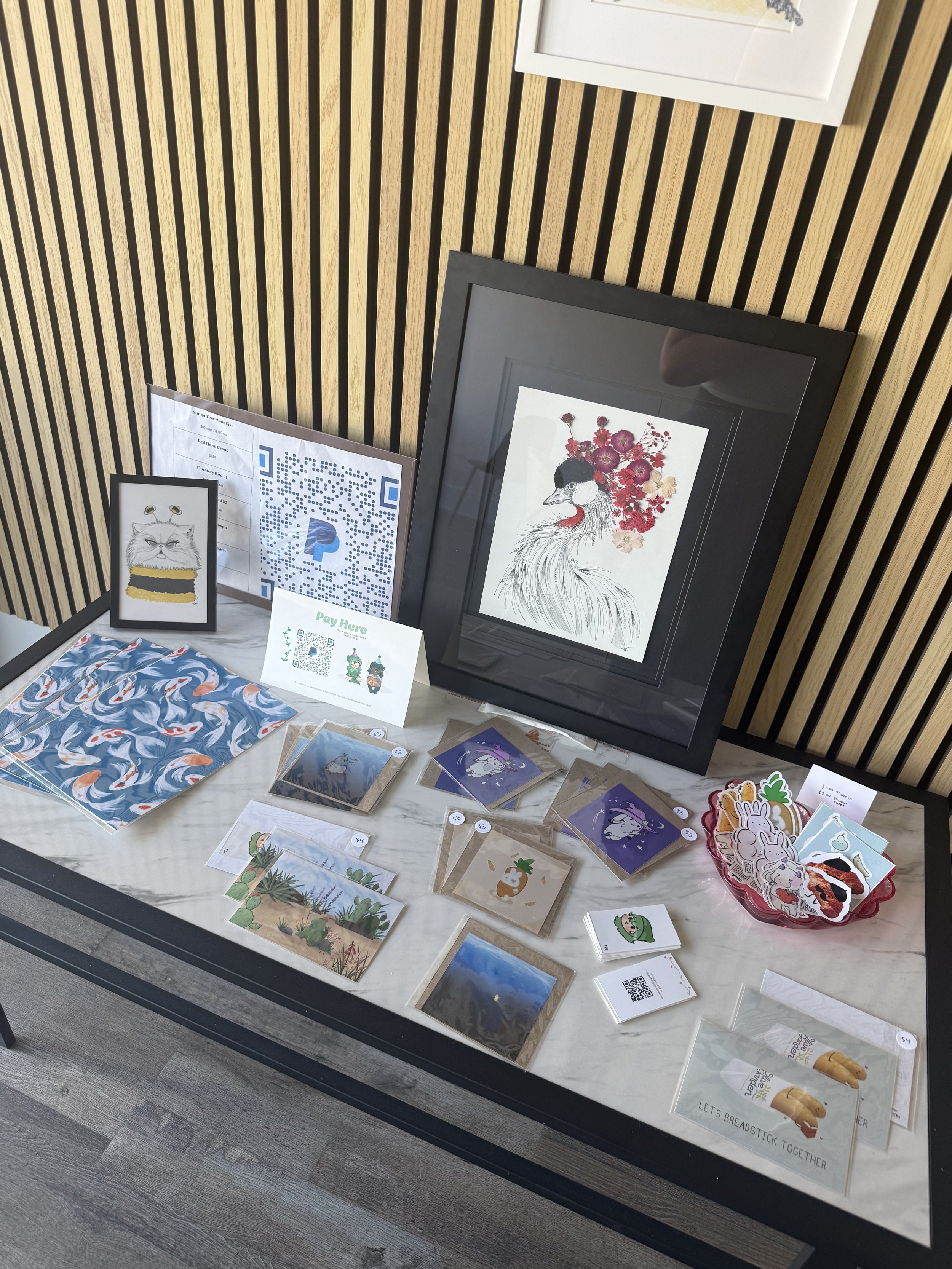 Display table showcasing framed artwork including a bird illustration and a cat illustration, along with postcards, stickers, QR codes, and a paper sign that says "Pay Here" in front of a wooden slatted wall.