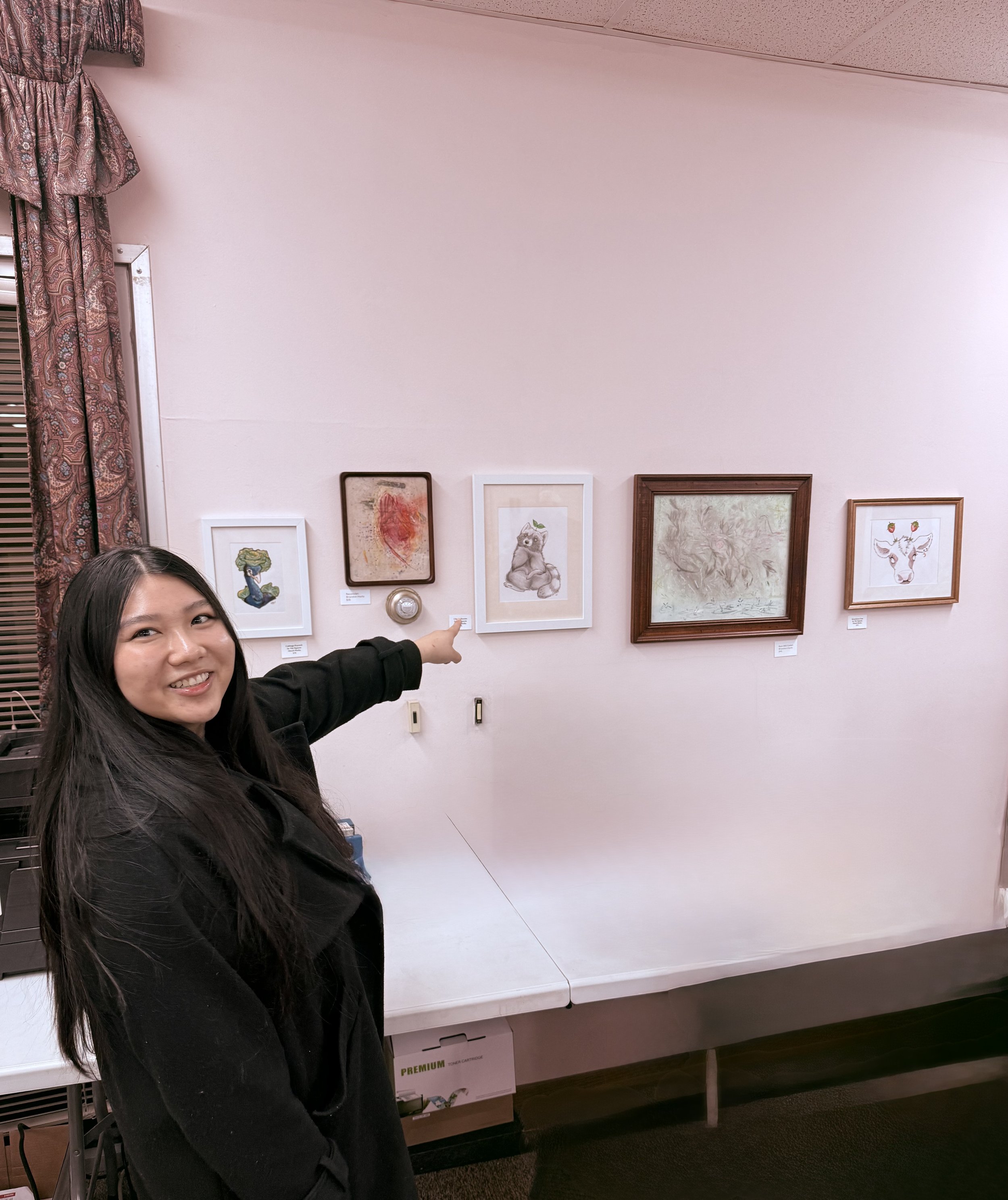 A woman pointing at framed artworks on a white wall. The artworks include a stylized animal, an abstract piece, a bear, a faint landscape, and a deer in a gallery setting.