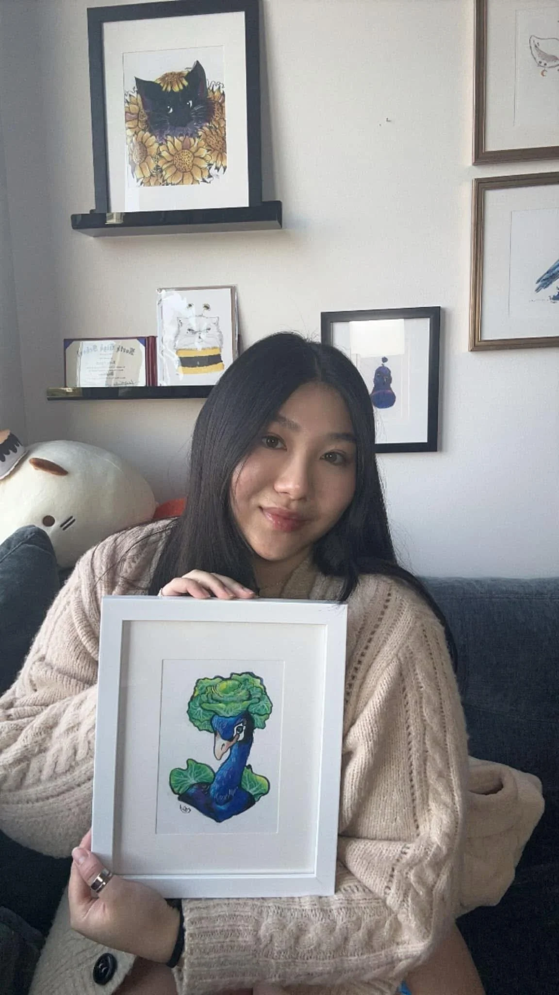 A woman with long dark hair sitting on a couch, holding a framed colorful illustration of a peacock with a head resembling a broccoli floret, in a cozy room decorated with framed artwork and plush toys.