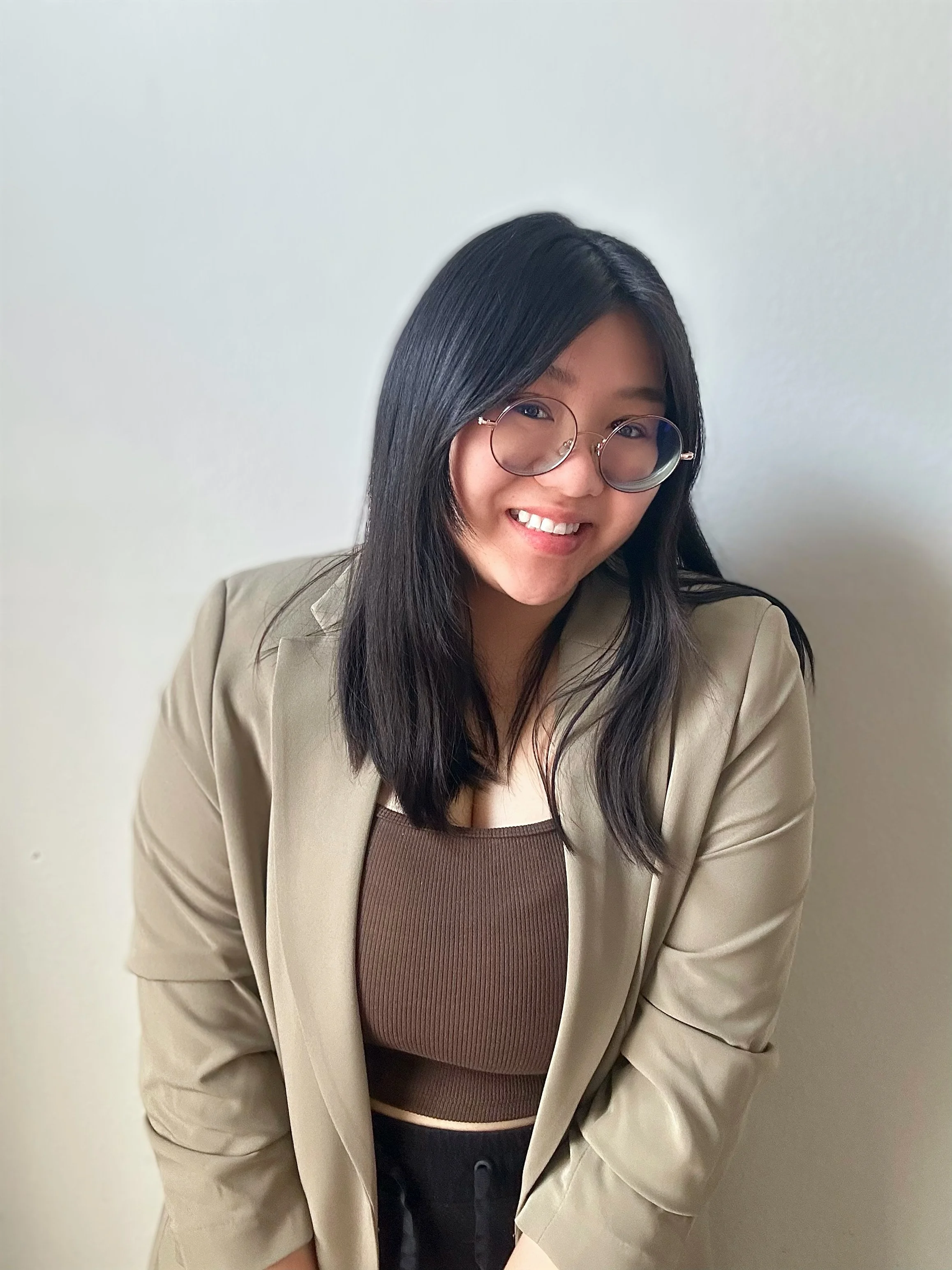 A young woman with shoulder-length black hair, wearing glasses, a beige blazer, a brown ribbed crop top, and black pants, smiling and leaning slightly forward against a plain white wall.