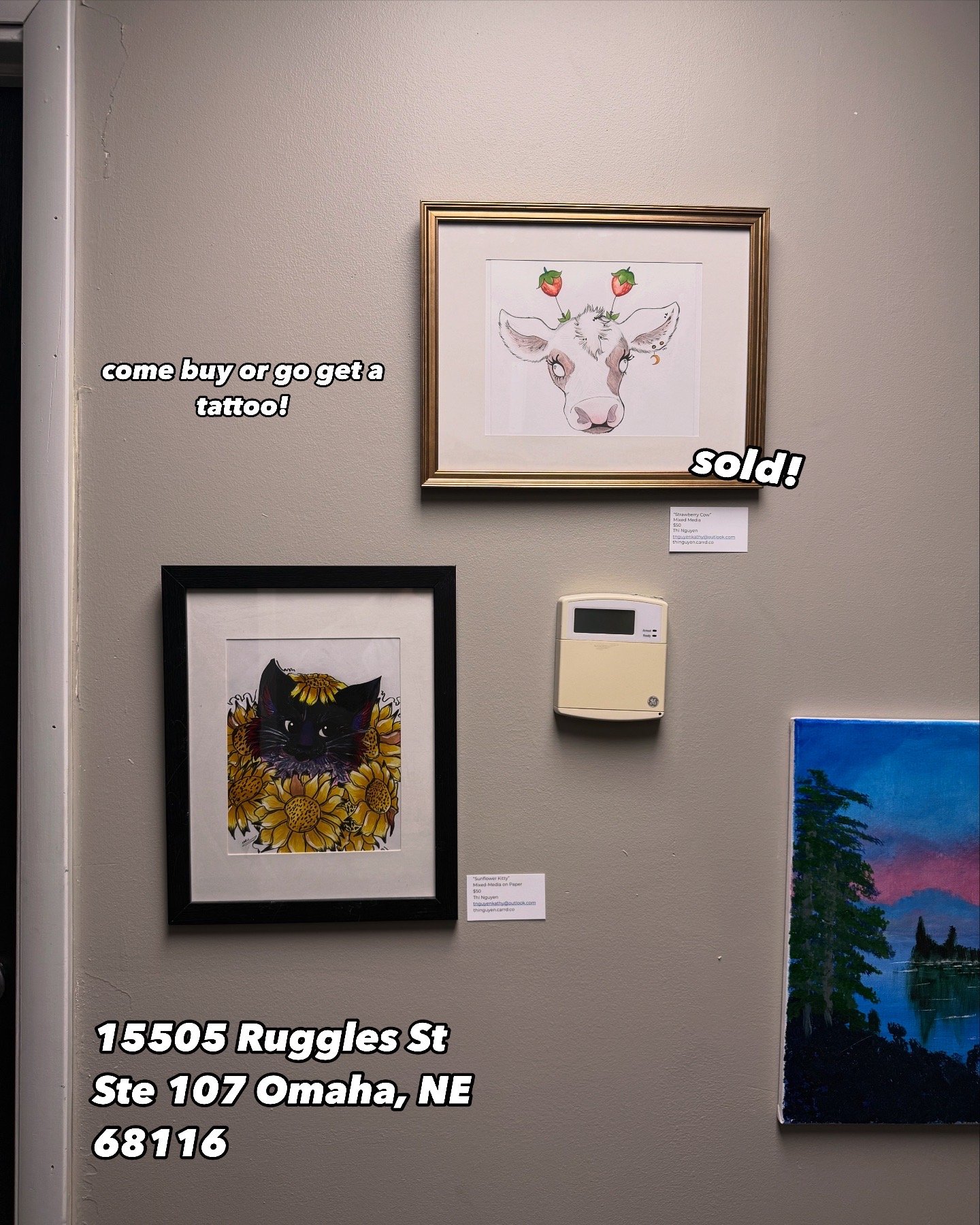 Art display with two framed watercolor paintings of animals, one of a cow and one of a cat, on a beige wall. The cow drawing has strawberries on its head, with a sign indicating it sold. The cat drawing features yellow flowers. Writing on the image p