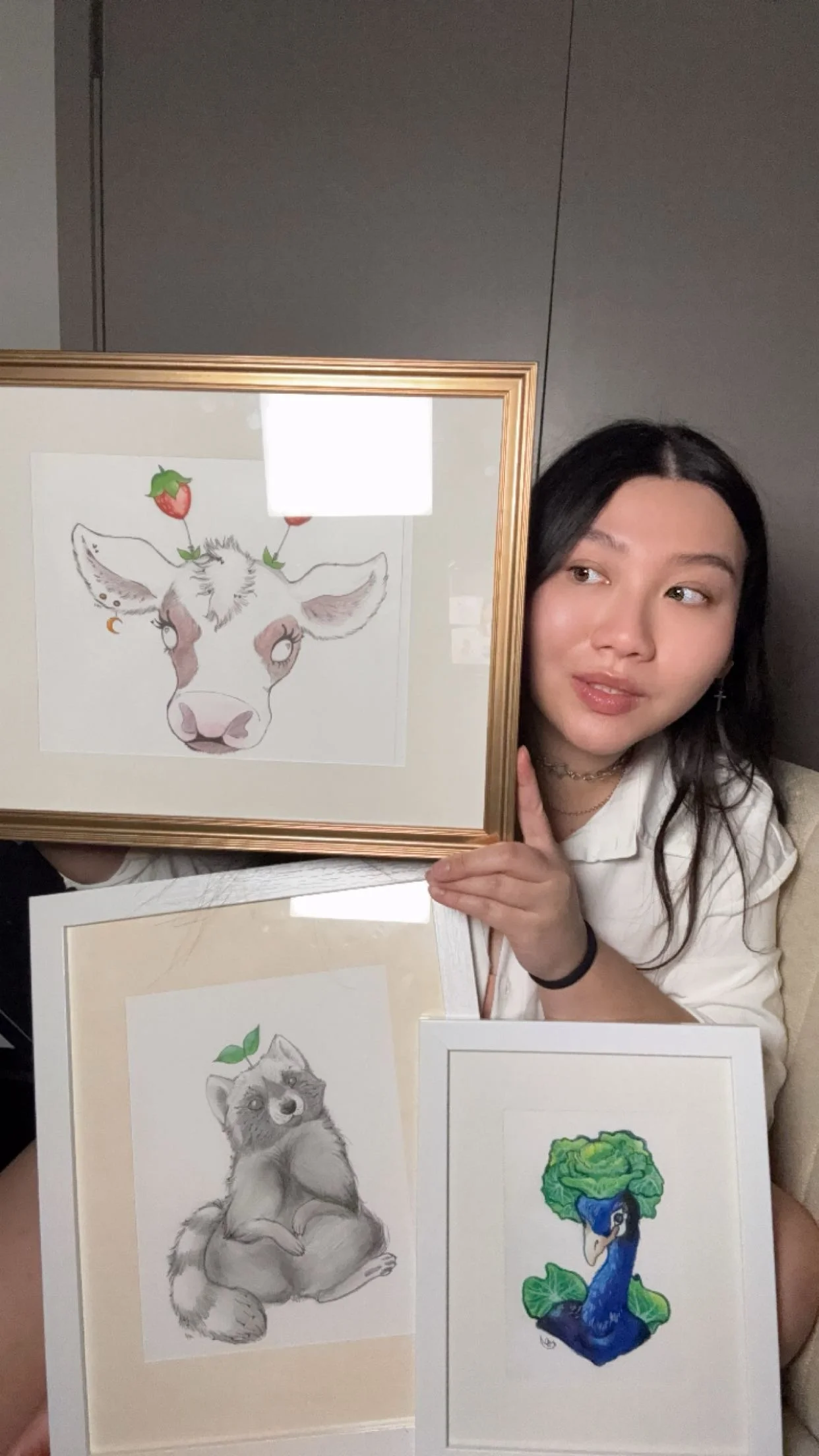 A woman with long black hair holding three framed animal illustrations, including a cow, a raccoon, and a peacock, in a room with dark walls.