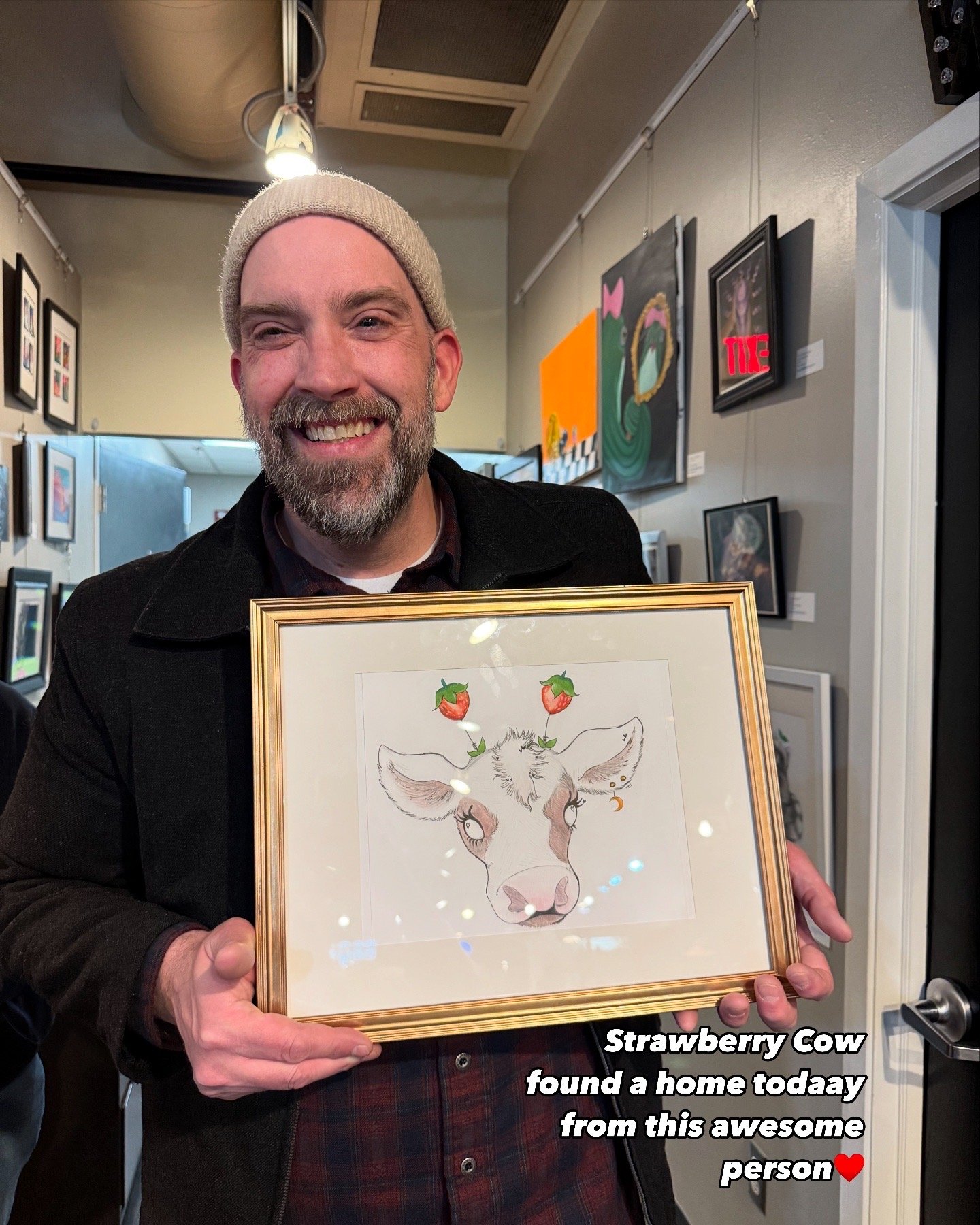 A man wearing a beige beanie and dark coat is smiling while holding a framed drawing of a cow with strawberries on its horns. The background shows art on the walls of an art gallery or studio.