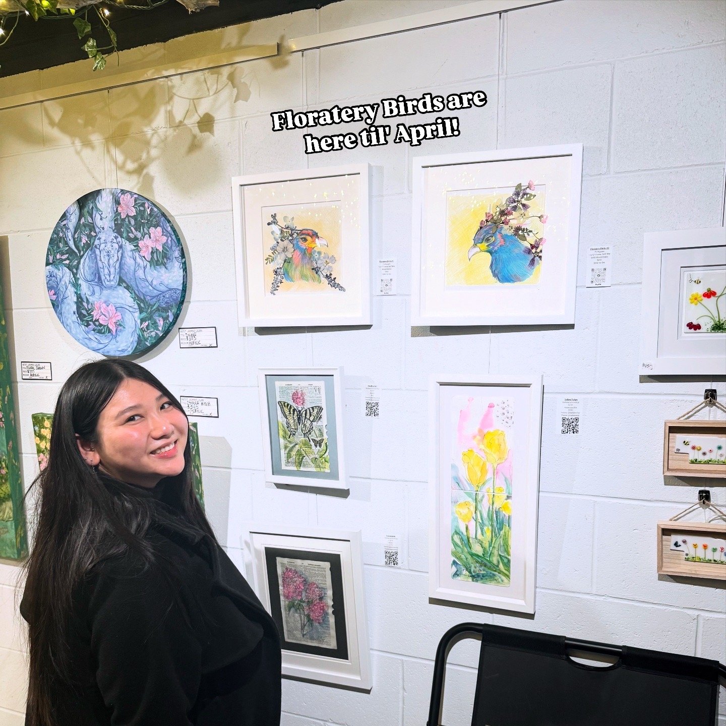 A woman with long dark hair and a black jacket smiling in front of a white wall with colorful bird and flower artwork. The wall has a sign that says 'Floratery Birds are here til April!'