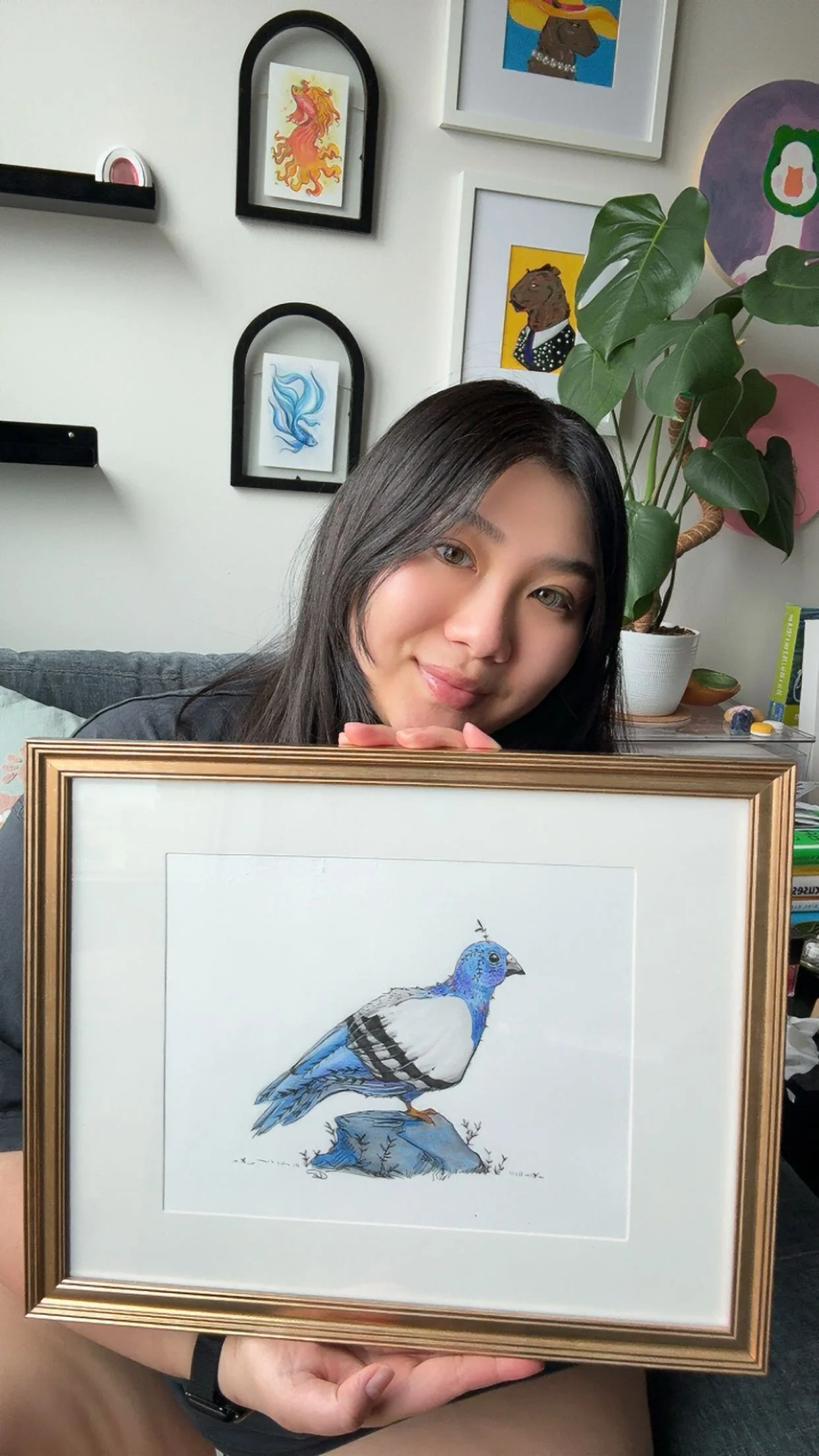 A woman with long dark hair smiling and holding a framed illustration of a blue and white bird perched on a rock.