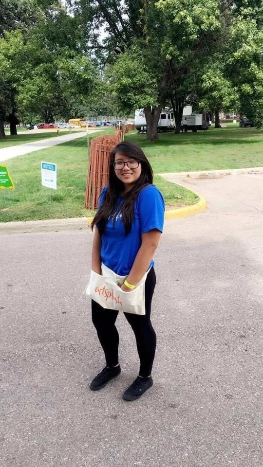 A young woman standing outdoors on a paved area, smiling at the camera. She has long dark hair, glasses, and wears a blue shirt, black pants, and black shoes. She carries a white bag with orange writing. Behind her are trees, a grassy area, a wooden 