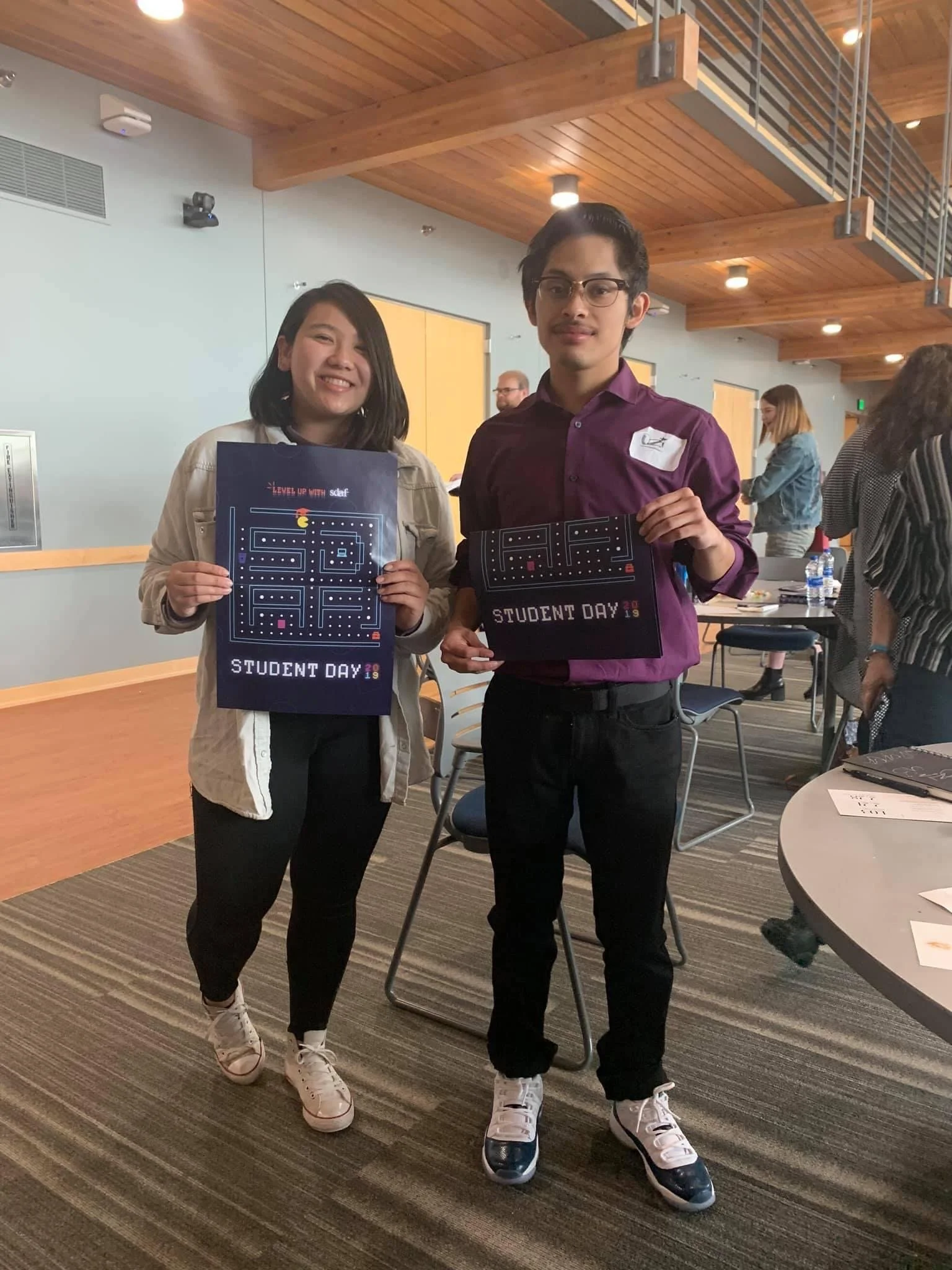 Two young people standing side by side, holding signs that read 'Student Day 2019.' The woman on the left has shoulder-length dark hair, is smiling, and wears a light-colored jacket and dark pants. The man on the right has glasses, short dark hair, a
