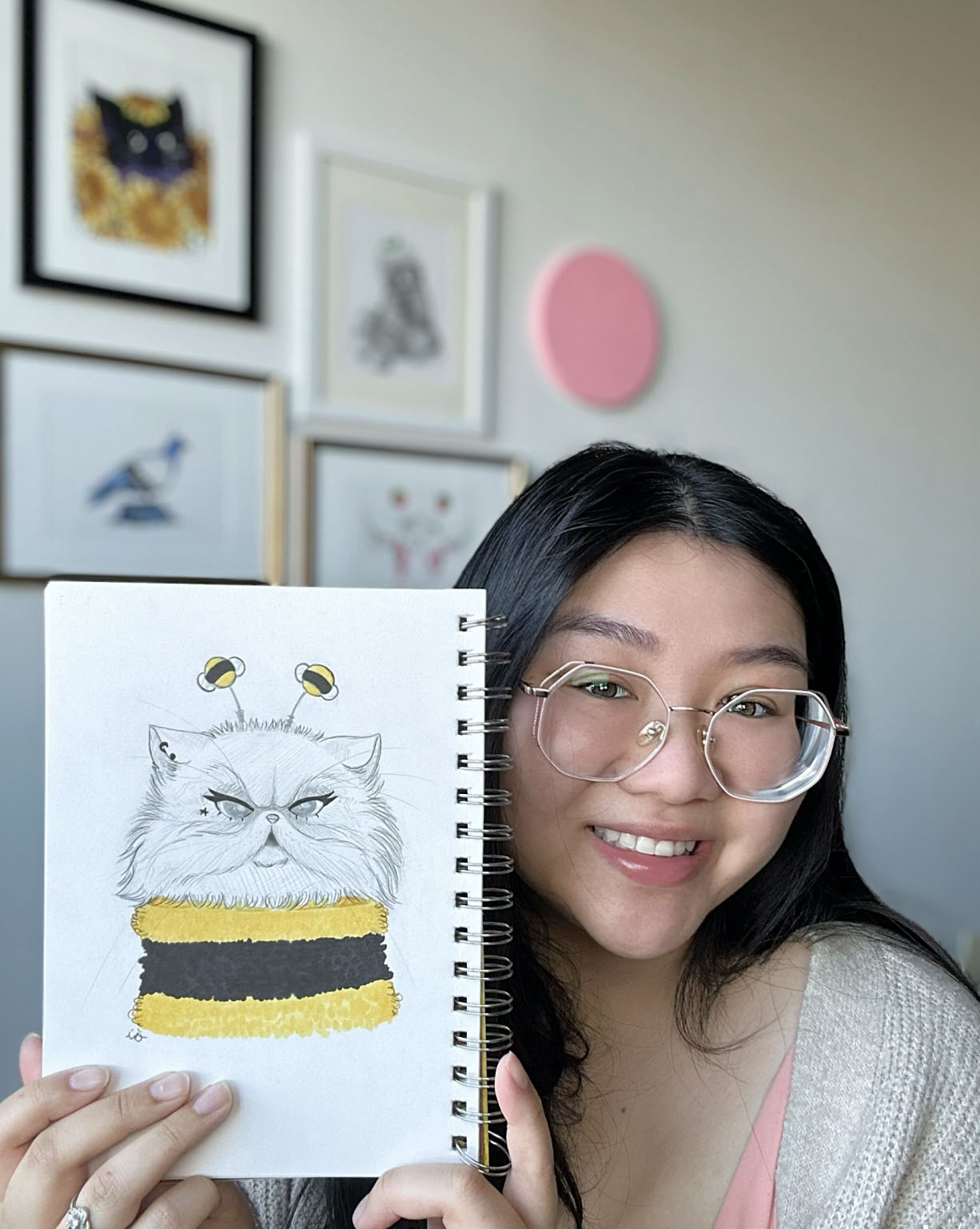 A young woman with glasses smiling and holding a sketchbook displaying a drawing of a grumpy cat with bee antennae and a yellow and black striped body.