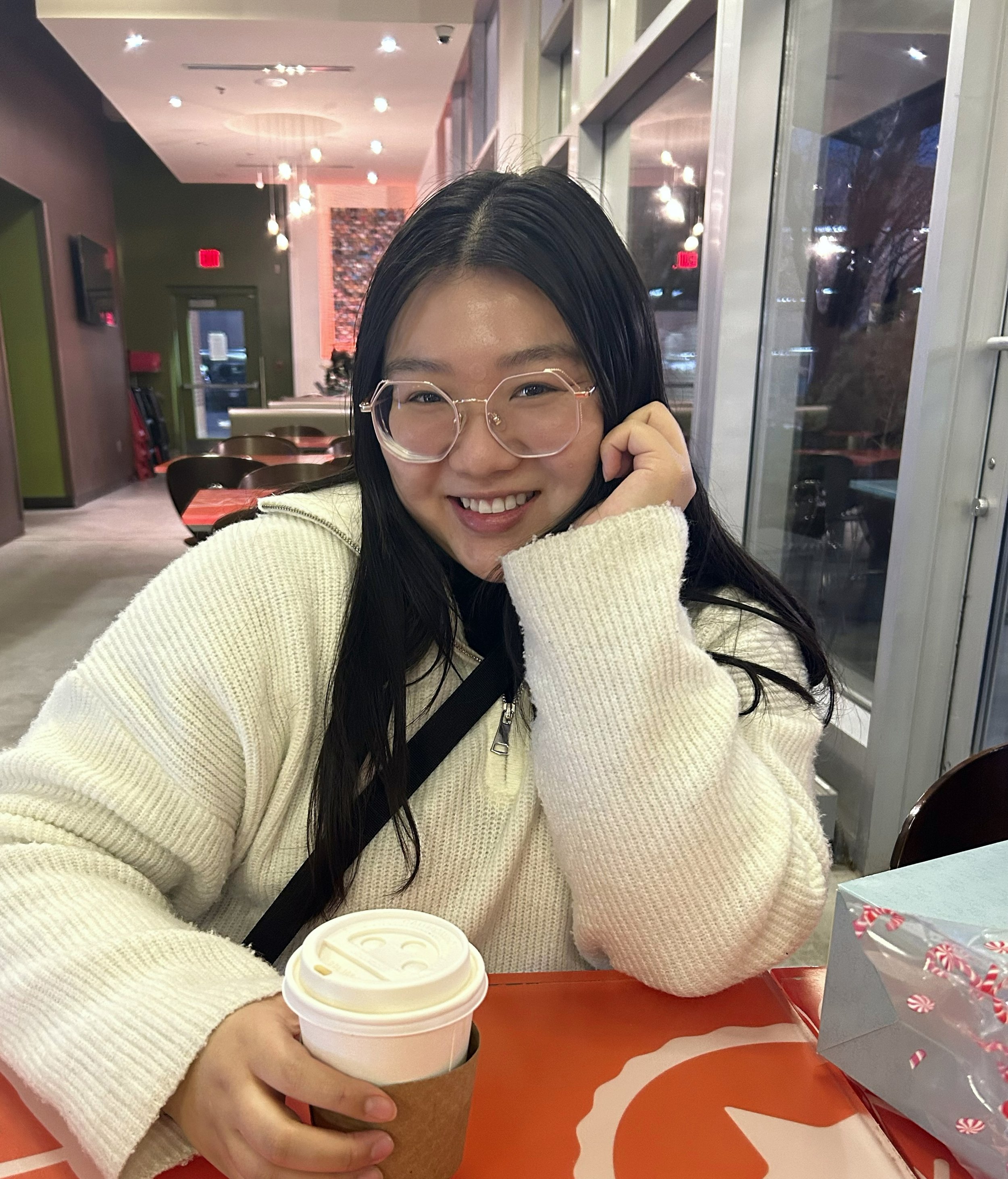 A young woman with long black hair, wearing glasses and a cream-colored sweater, smiling and sitting at a table with a warm drink in a festive, brightly lit cafe.