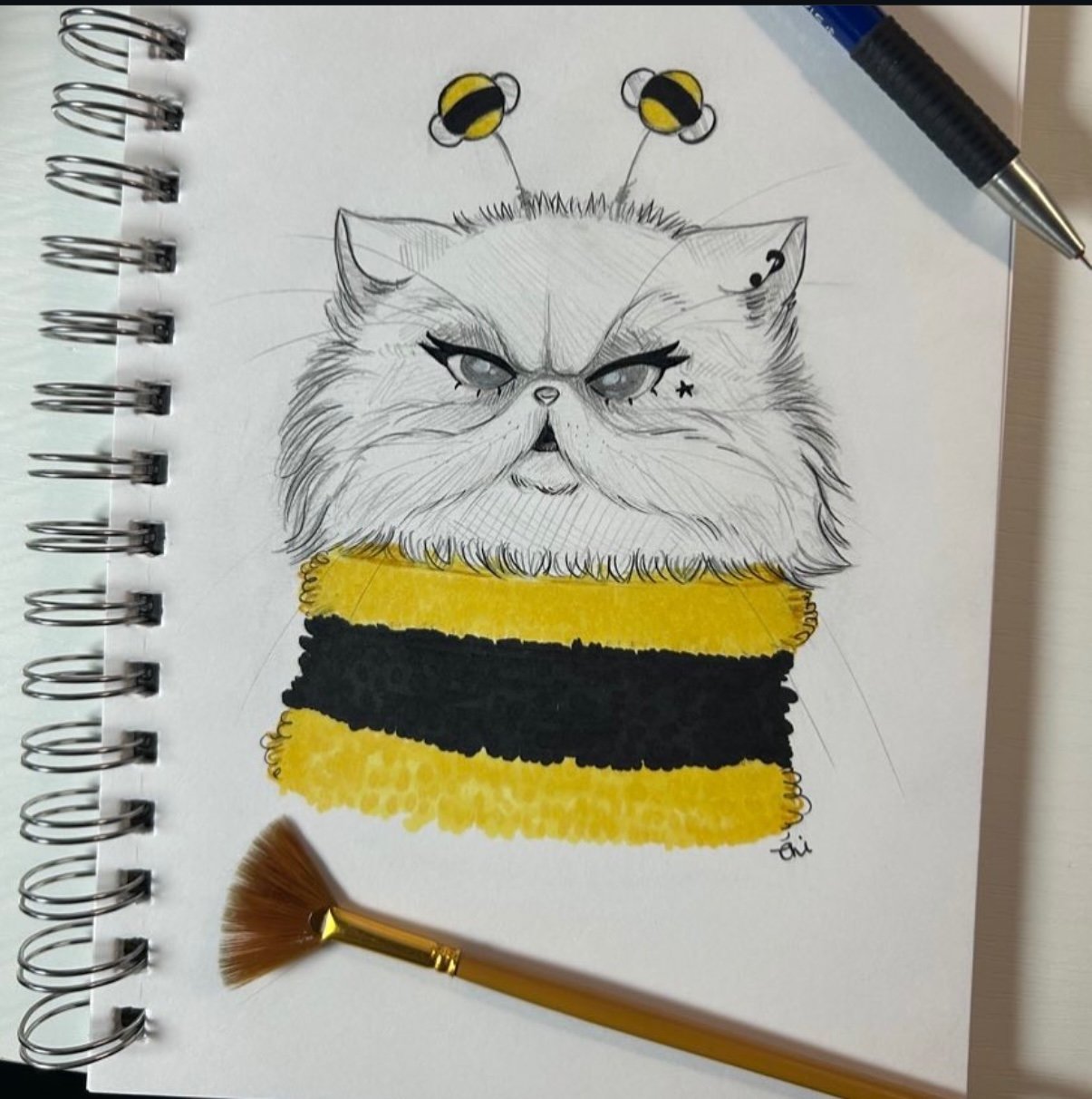 A sketch of a grumpy cat with gray eyes, black eyeliner, and a small star tattoo under one eye, wearing a yellow and black striped sweater, with bee antennae on its head, and a fan brush placed below the drawing. The sketch is on a spiral notebook wi