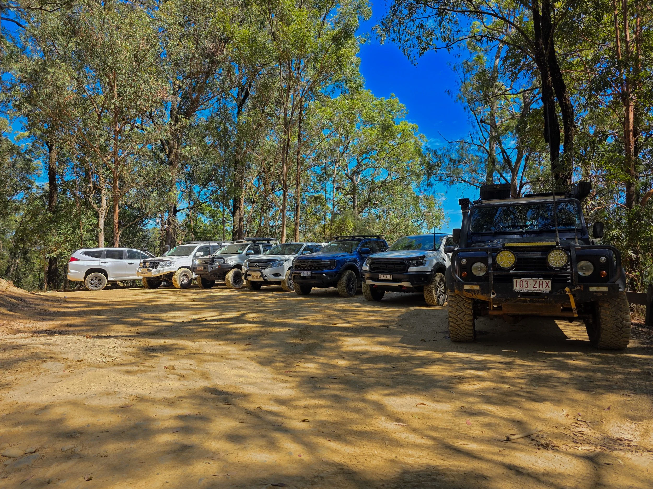 Several off-road vehicles, including SUVs and a rugged all-terrain vehicle, parked on a dirt clearing among trees with green leaves and a bright blue sky overhead.