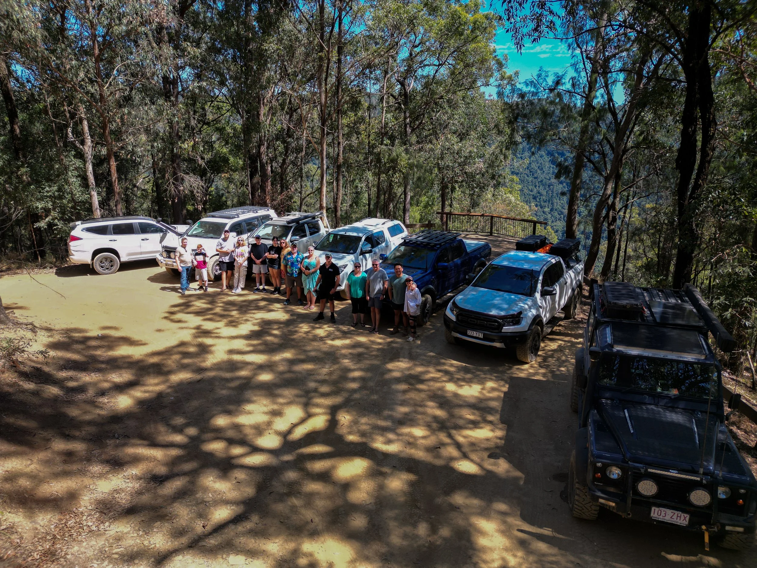 Group of people standing in a line in front of parked off-road vehicles on a dirt clearing in a forest, with trees and a mountain view in the background.