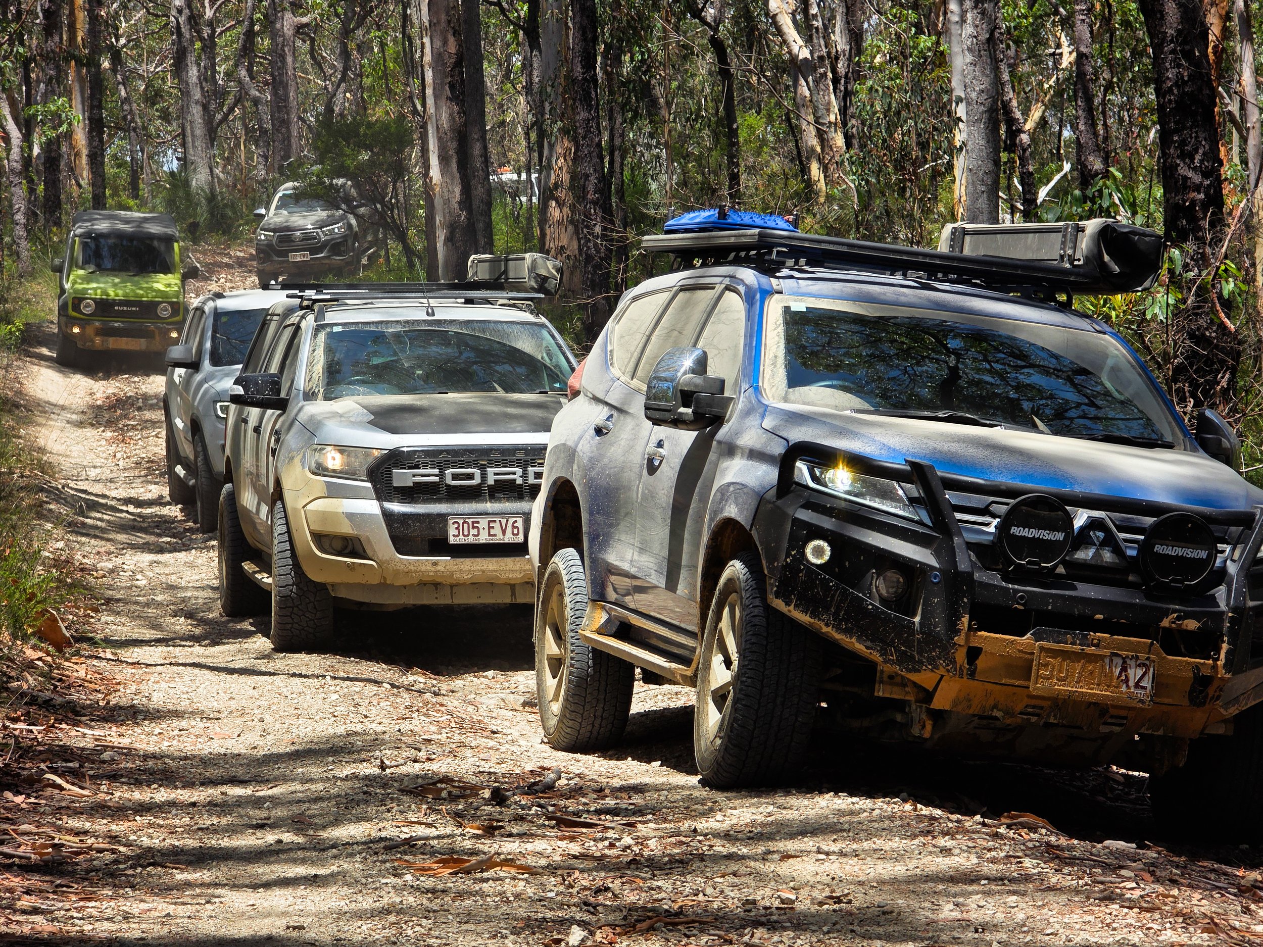 Off-road cars driving through a forest trail, with dust and dirt on their tires and bodywork, surrounded by trees and foliage.