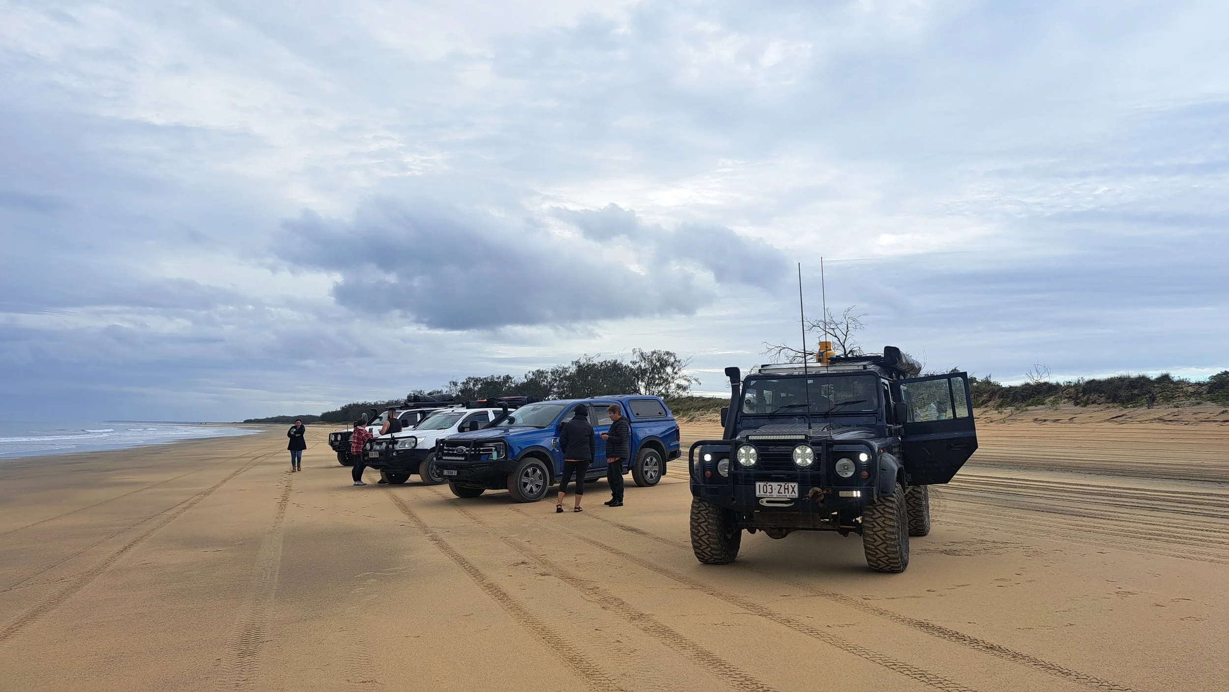 Five off-road vehicles parked on a sandy beach with a cloudy sky. People are standing near the vehicles, some talking, one person walking further away. Tire tracks are visible in the sand, and the ocean is to the left.