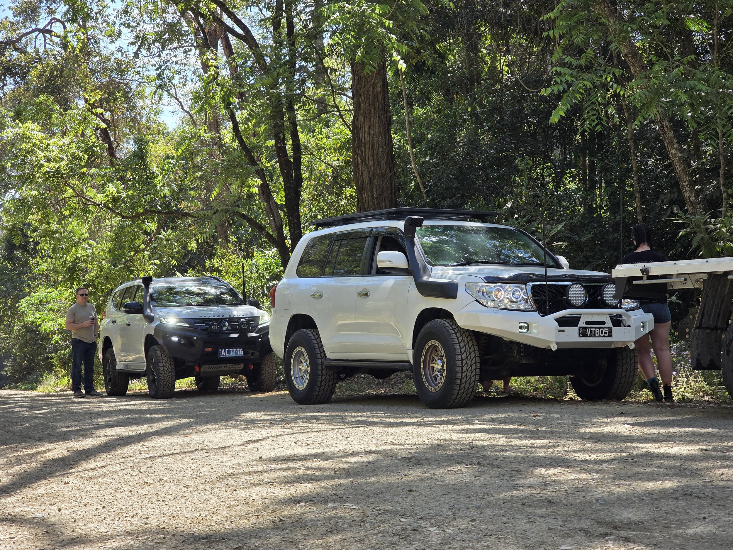 Two off-road vehicles parked on a dirt trail in a forest, with a man and a woman nearby, surrounded by green trees and sunlight filtering through the leaves.