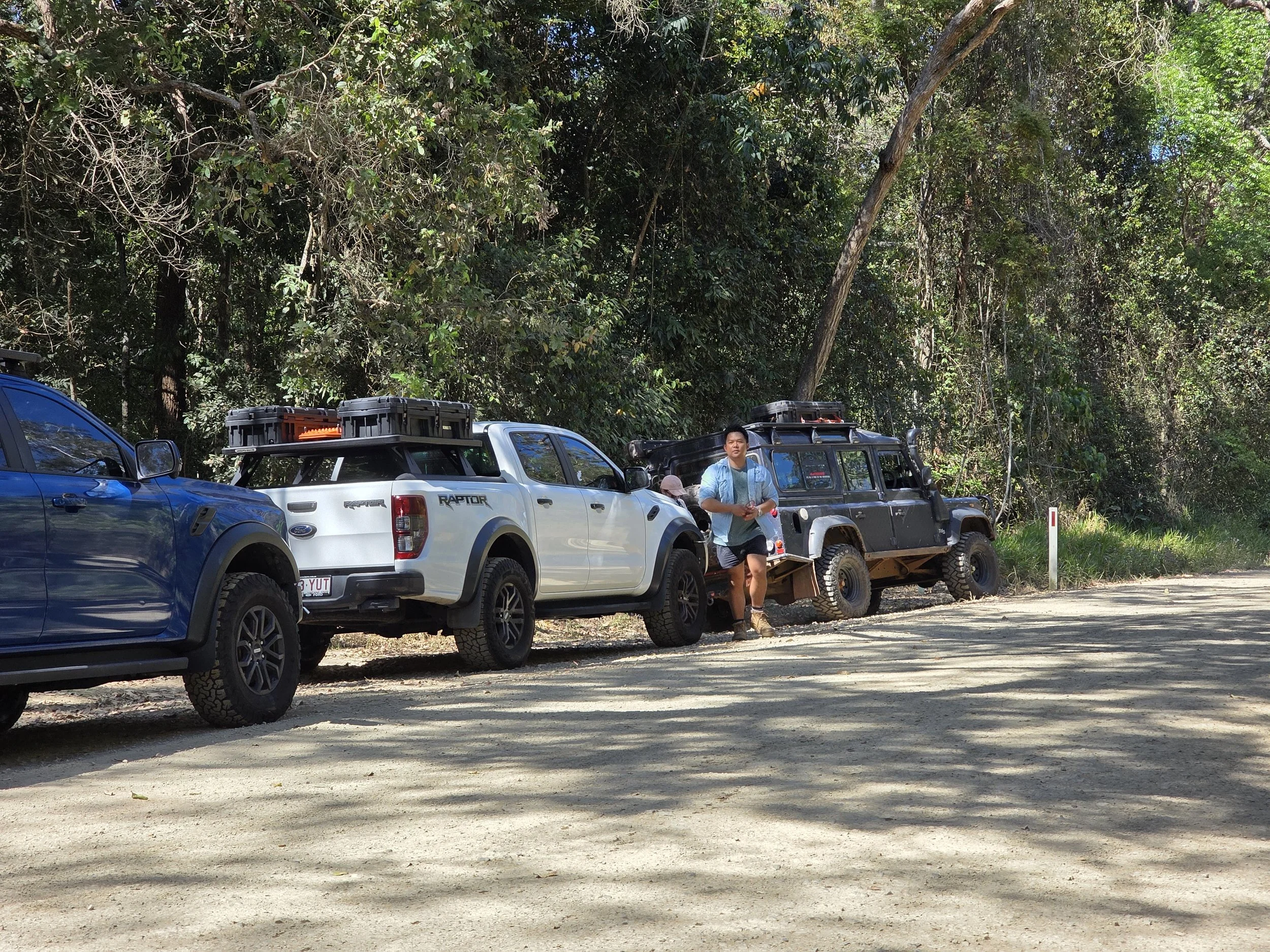 Three parked off-road vehicles on a dirt road next to a person standing among them, with a dense forest in the background.