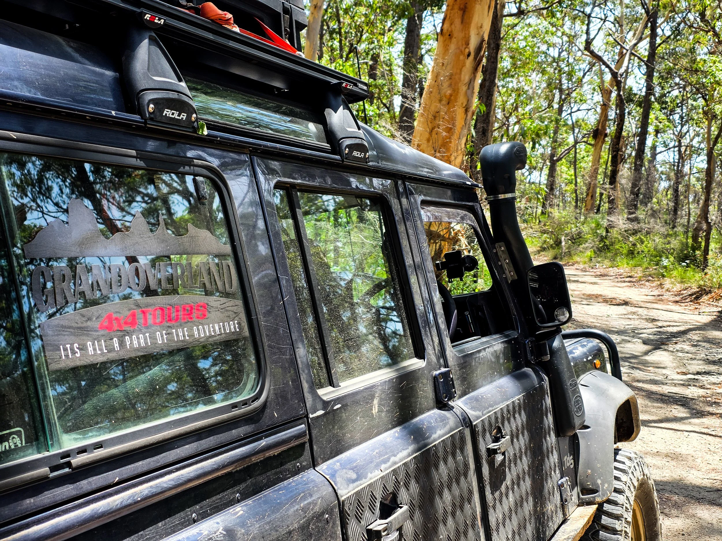 Close-up of a black off-road vehicle on a dirt trail surrounded by trees, with a sticker on the window that reads 'GRANDOVERLAND 4x4 TOURS' and 'ITS ALL A PART OF THE ADVENTURE'.