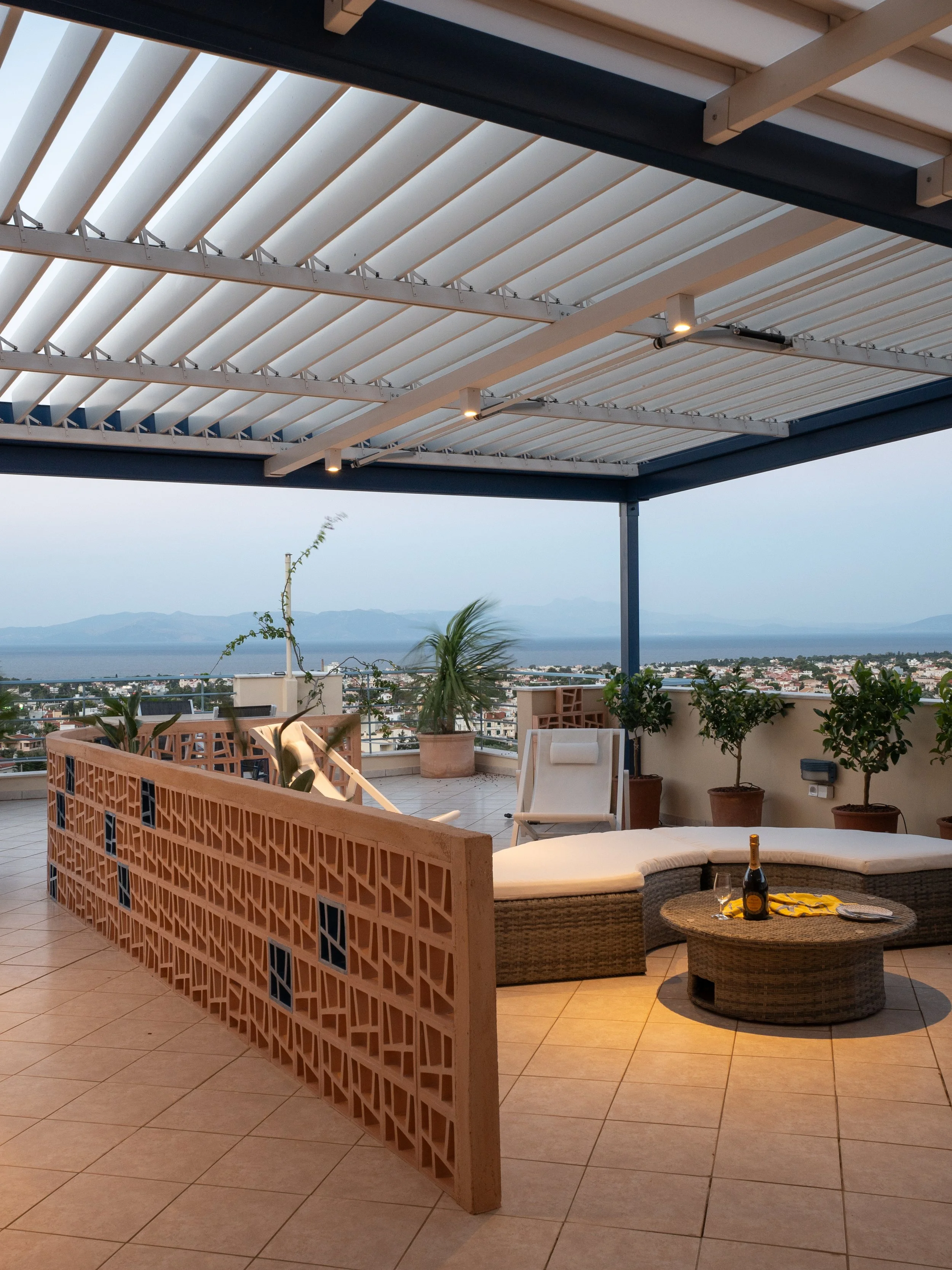 An outdoor rooftop patio area with a view of the city and mountains in the background. The space includes a decorative brick railing, potted plants, a seating area with a round wicker table, lounge chairs, and a bottle of champagne with glasses on th