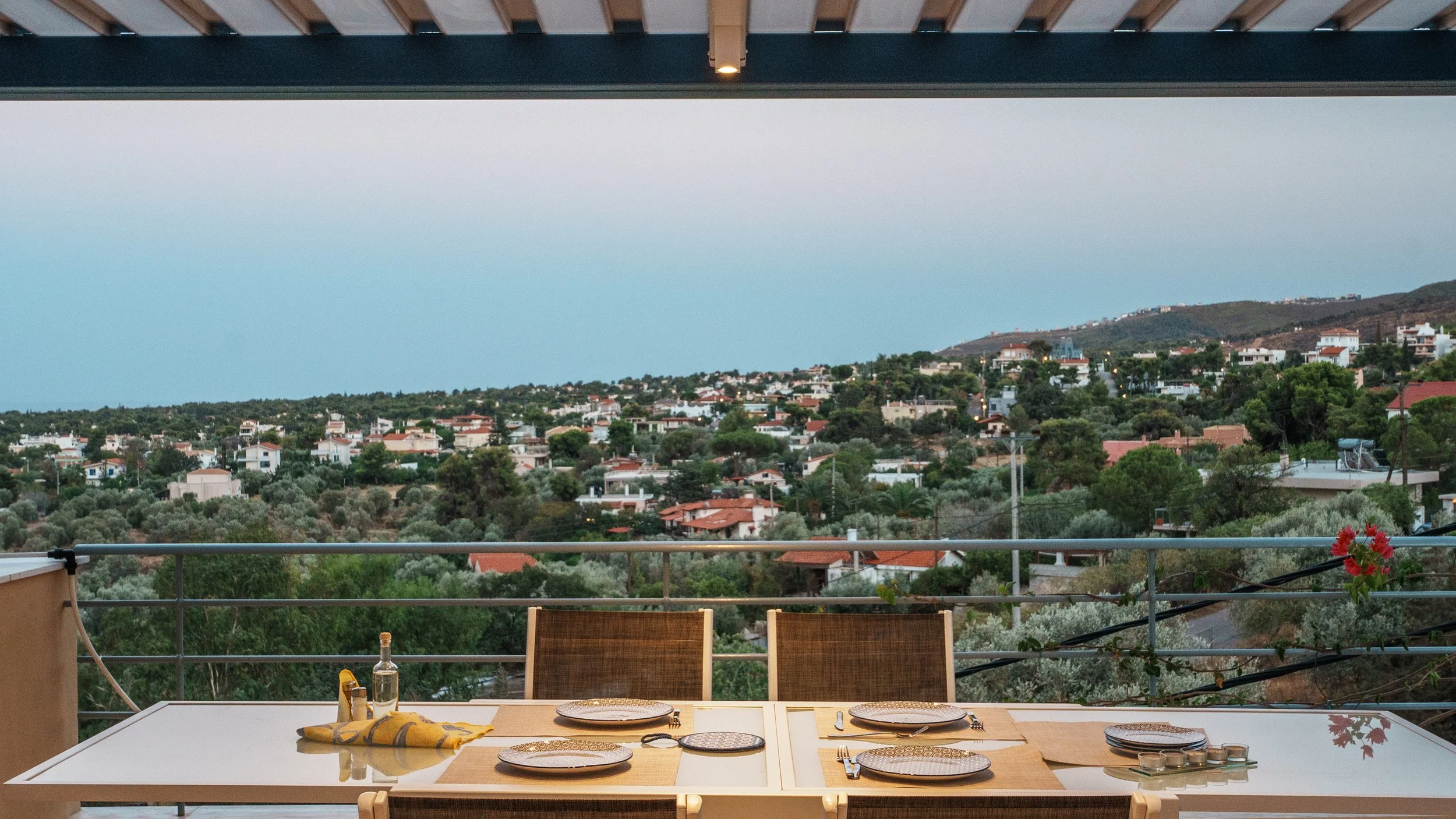 A dining table with placemats, plates, and silverware on a balcony overlooking a residential neighborhood with trees and hills during dusk.