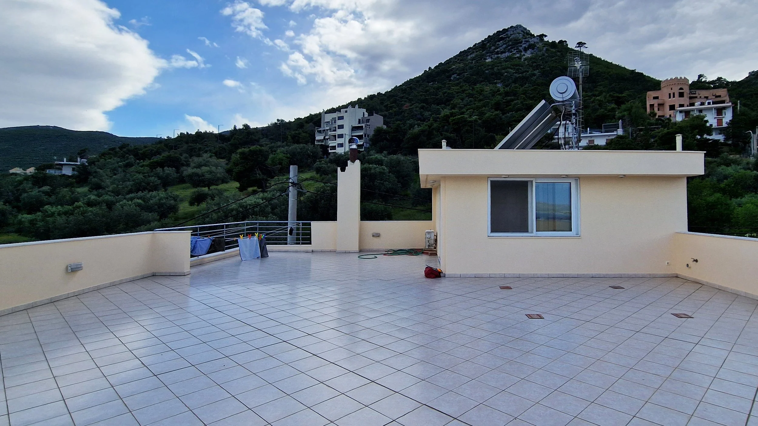 A spacious outdoor terrace with beige tiled flooring, a small building with a window, laundry hanging on a clothesline, and a hillside with trees and residential buildings in the background under a partly cloudy sky.