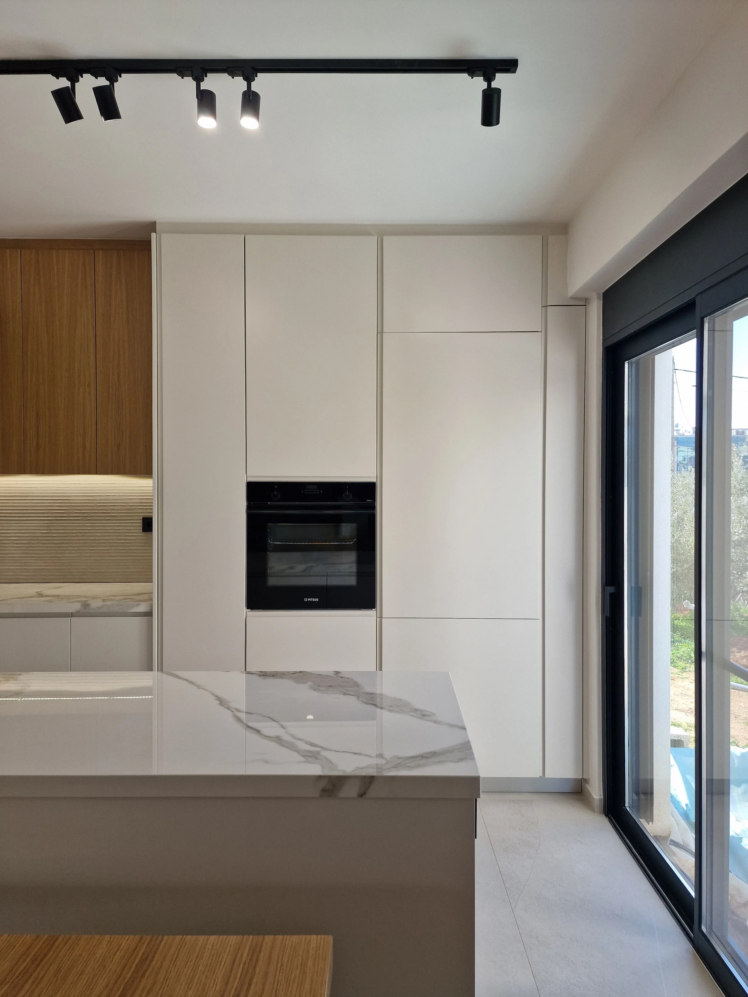 Modern kitchen with white marble island, built-in oven, white cabinets, wooden accent wall, sliding glass door, and track lighting on the ceiling.