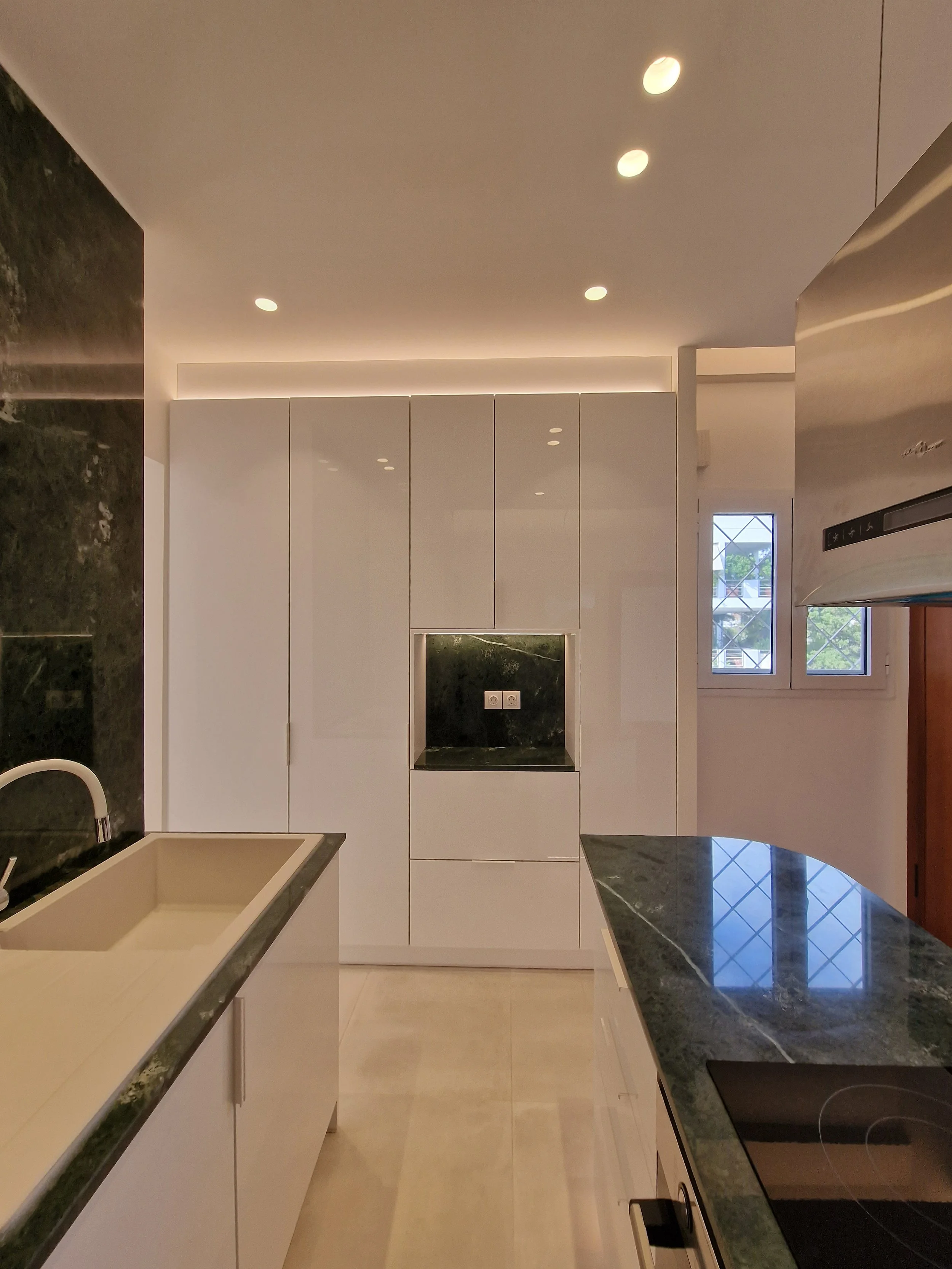 A modern kitchen with white cabinets, green marble countertops, a sink, and a small window.