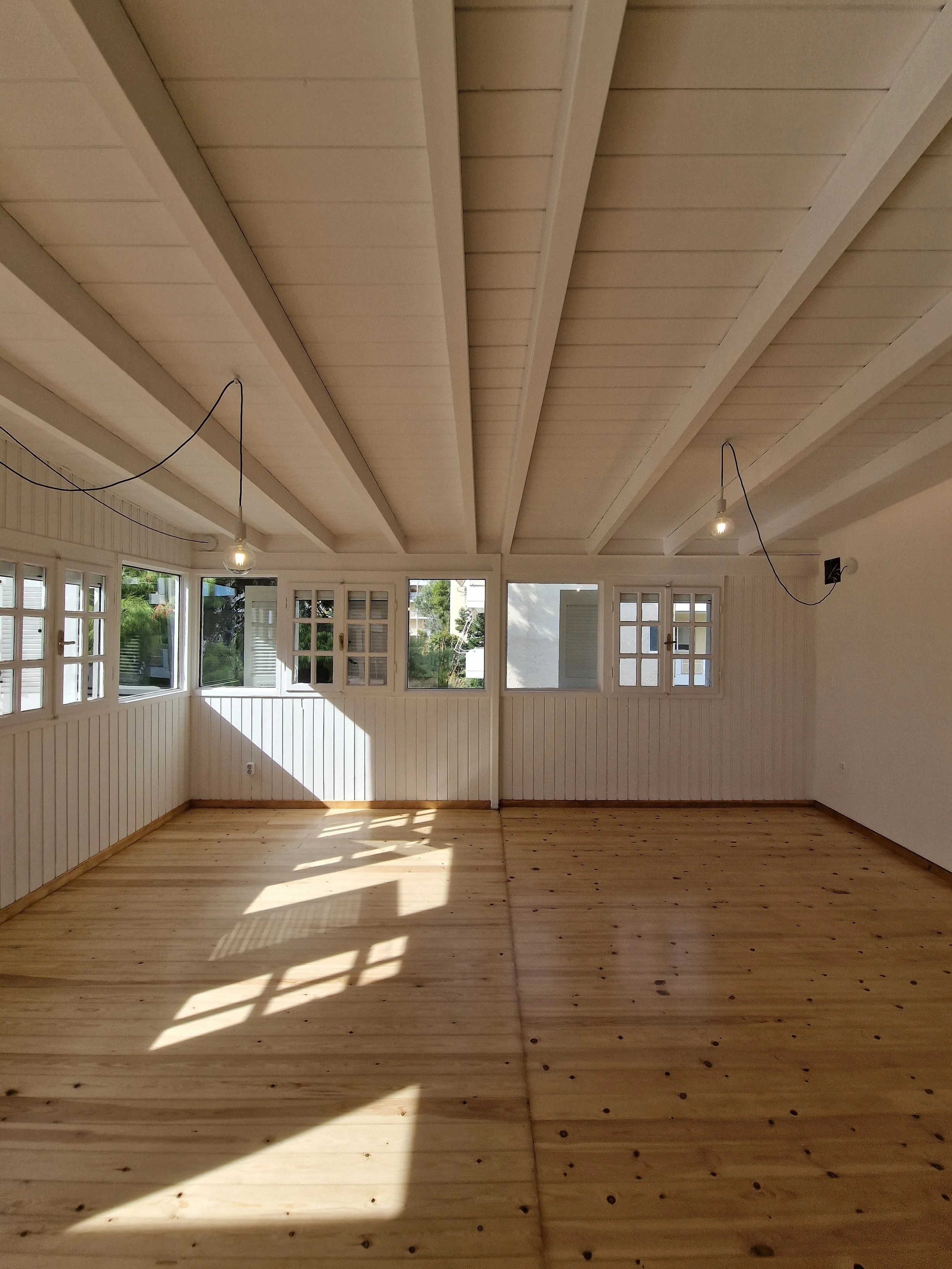 Empty room with wooden flooring, white paneled walls, multiple windows, and exposed light bulbs hanging from the ceiling.