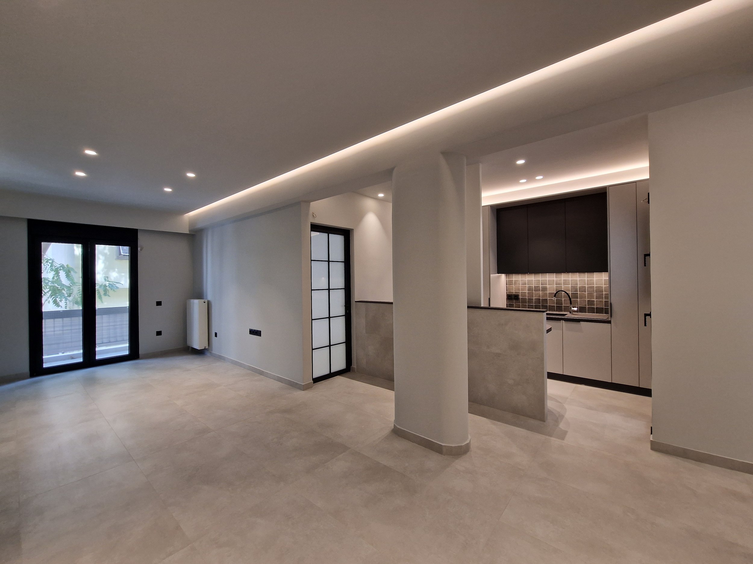 Modern open-plan living area with large tiled floor, white walls, multiple ceiling lights, black framed glass door leading to an outdoor patio, and a kitchen area with black cabinets and a tiled backsplash.