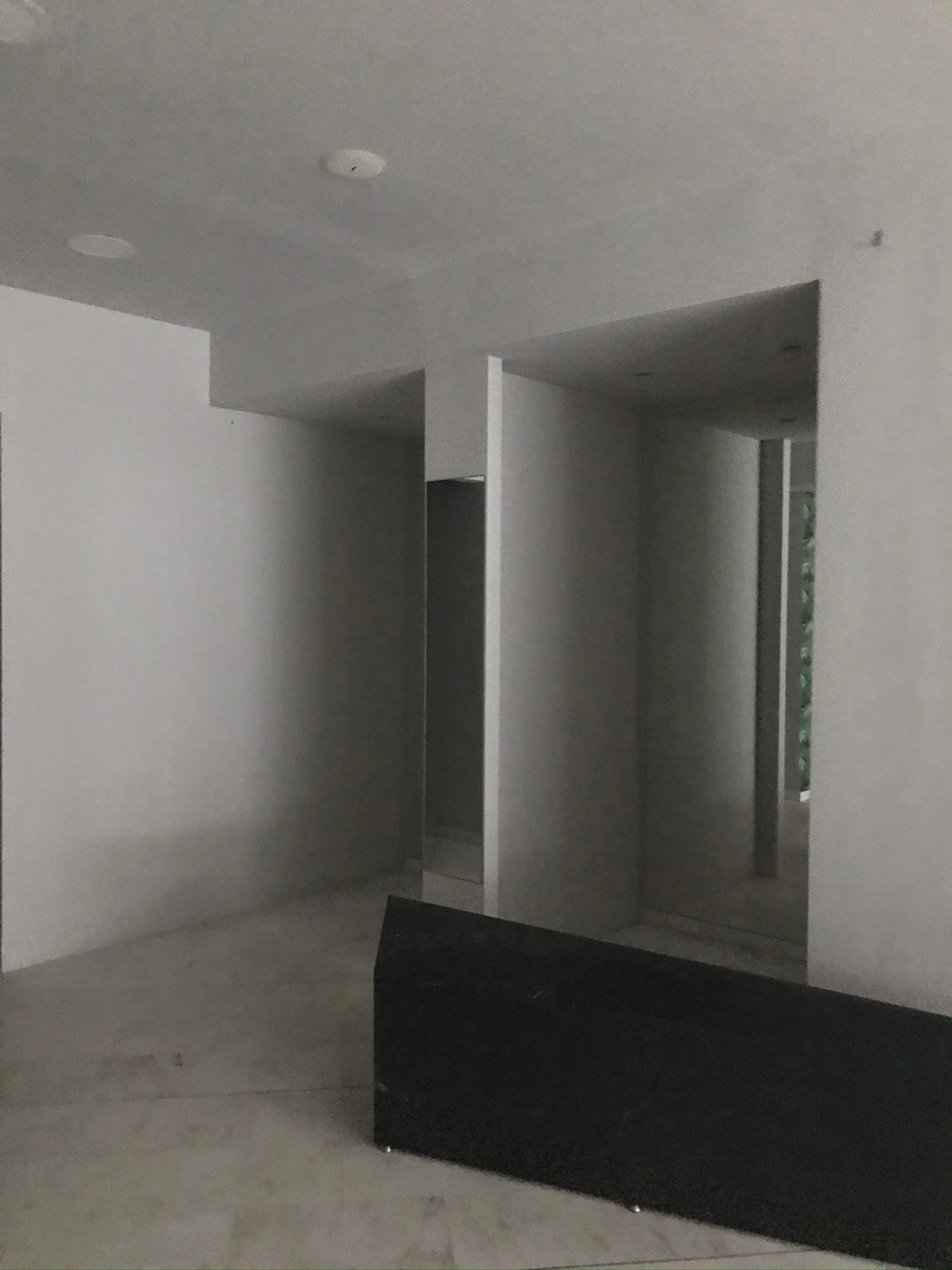 Empty interior space of a modern house with white walls, a ceiling with recessed lighting, and a black piece of furniture or structure near a doorway.