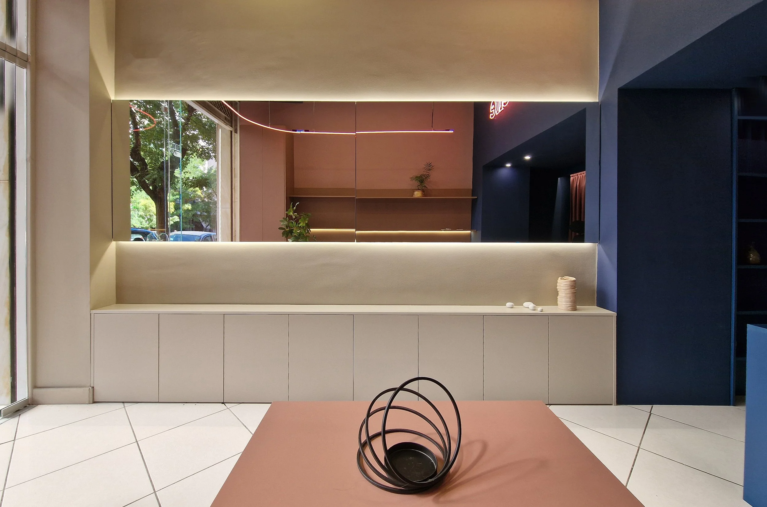 Modern interior space with a large mirror, potted plants, a pink wall, a window with outside view, and a table with a decorative object. Part of a seating area with a neon sign reading 'SLEEP' is visible.