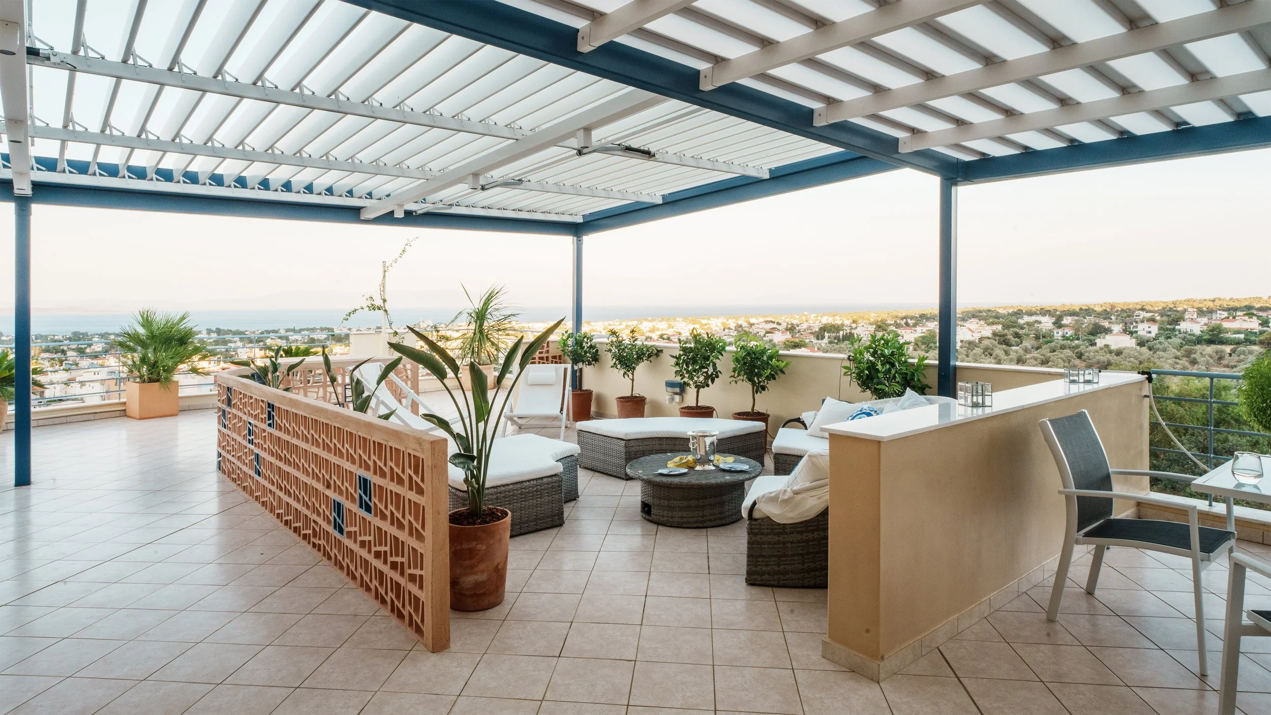 A spacious rooftop terrace with outdoor seating, including wicker chairs with white cushions and a small round table set with drinks and snacks. The terrace is decorated with potted plants and has a view of a cityscape and distant water horizon, with