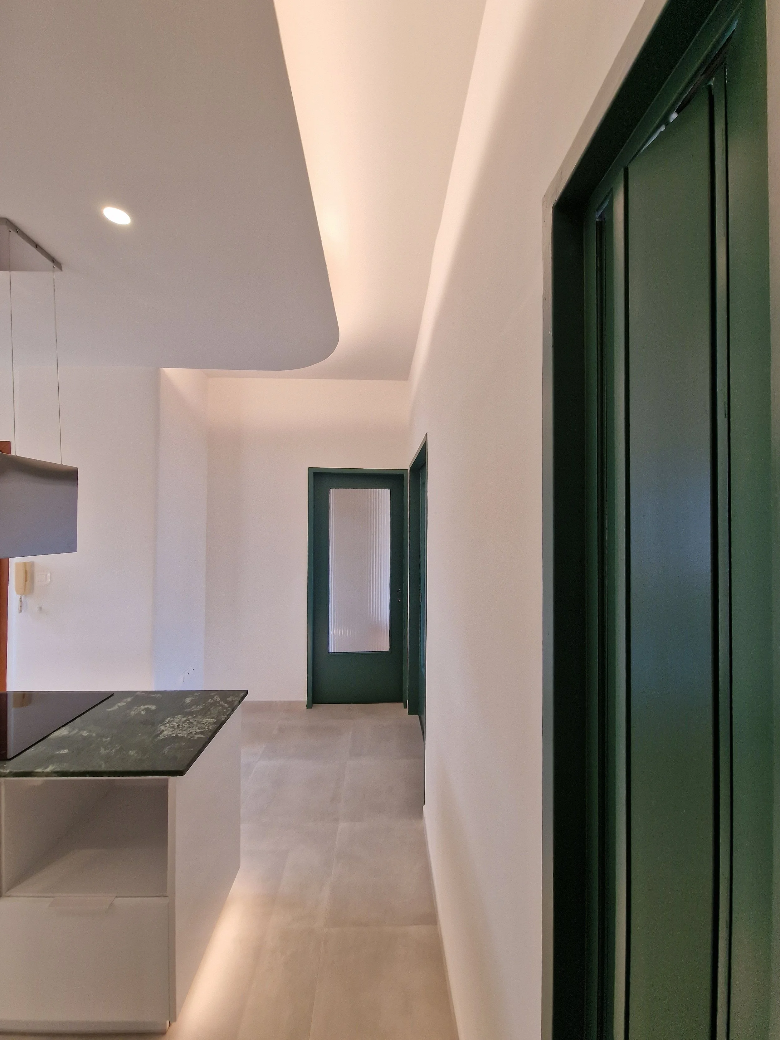 An interior hallway with white walls, beige tiled floor, green elevator doors on the right side, and a door at the end of the hallway.