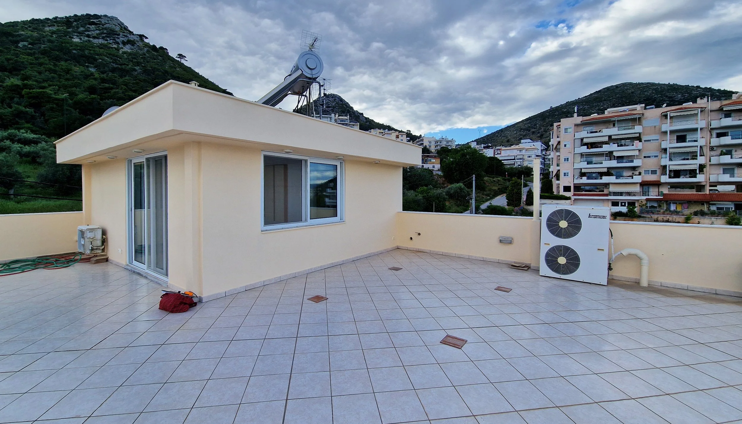 A rooftop terrace with tiled flooring, light-colored walls, a window, and sliding glass door. There is an outdoor air conditioning unit, some hoses, and a red bag. In the background, there are hills with trees and multi-story residential buildings, u