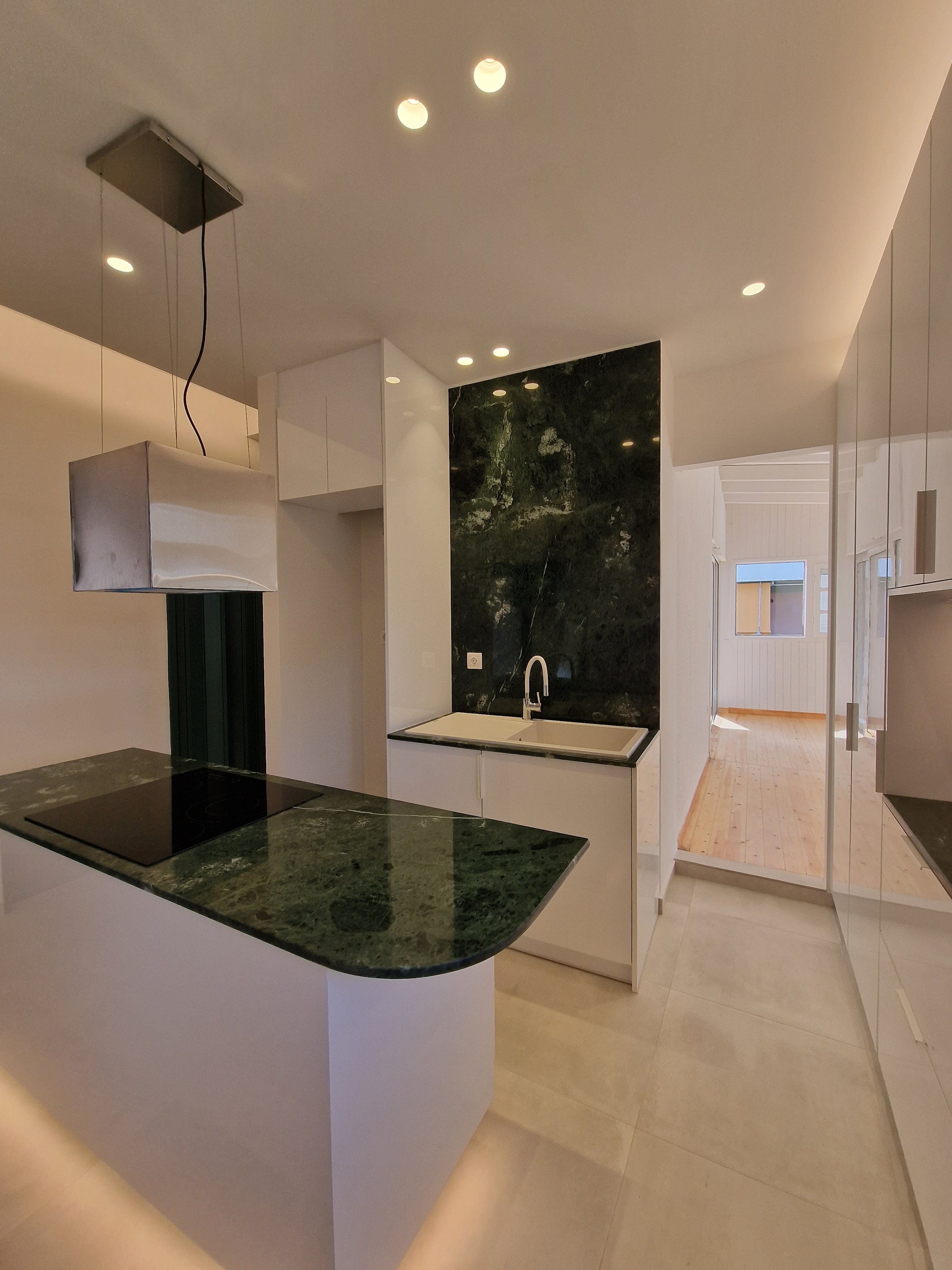 A modern kitchen with a dark green marble countertop, white cabinets, and a black induction stove. There is a large dark green marble backsplash behind the sink. In the background, there is a doorway leading to a well-lit room with wooden flooring an
