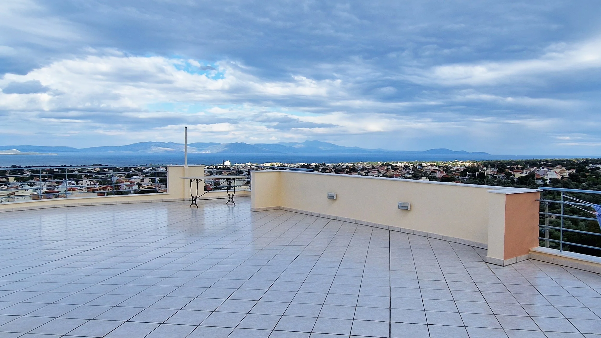 A rooftop terrace with tiled flooring and a beige railing overlooking a town, with mountains and a body of water in the distance under a partly cloudy sky.