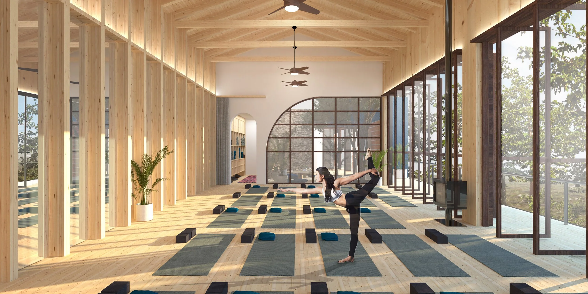 A woman practicing yoga in a bright, modern wooden-floored yoga studio with large windows, ceiling fans, and a woman in a side plank pose on a mat.