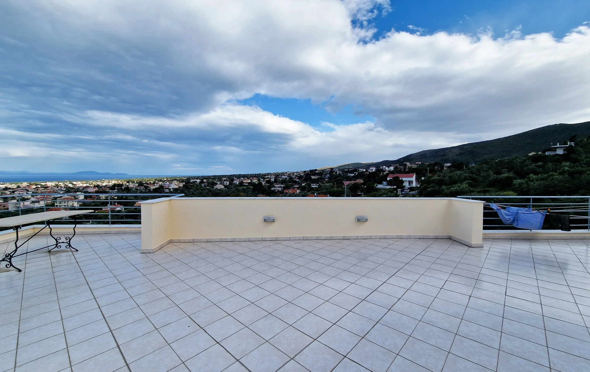 A rooftop terrace with tiled flooring, a clothesline with a blue cloth, a table, and a view of a hilly landscape with houses under a partly cloudy sky.