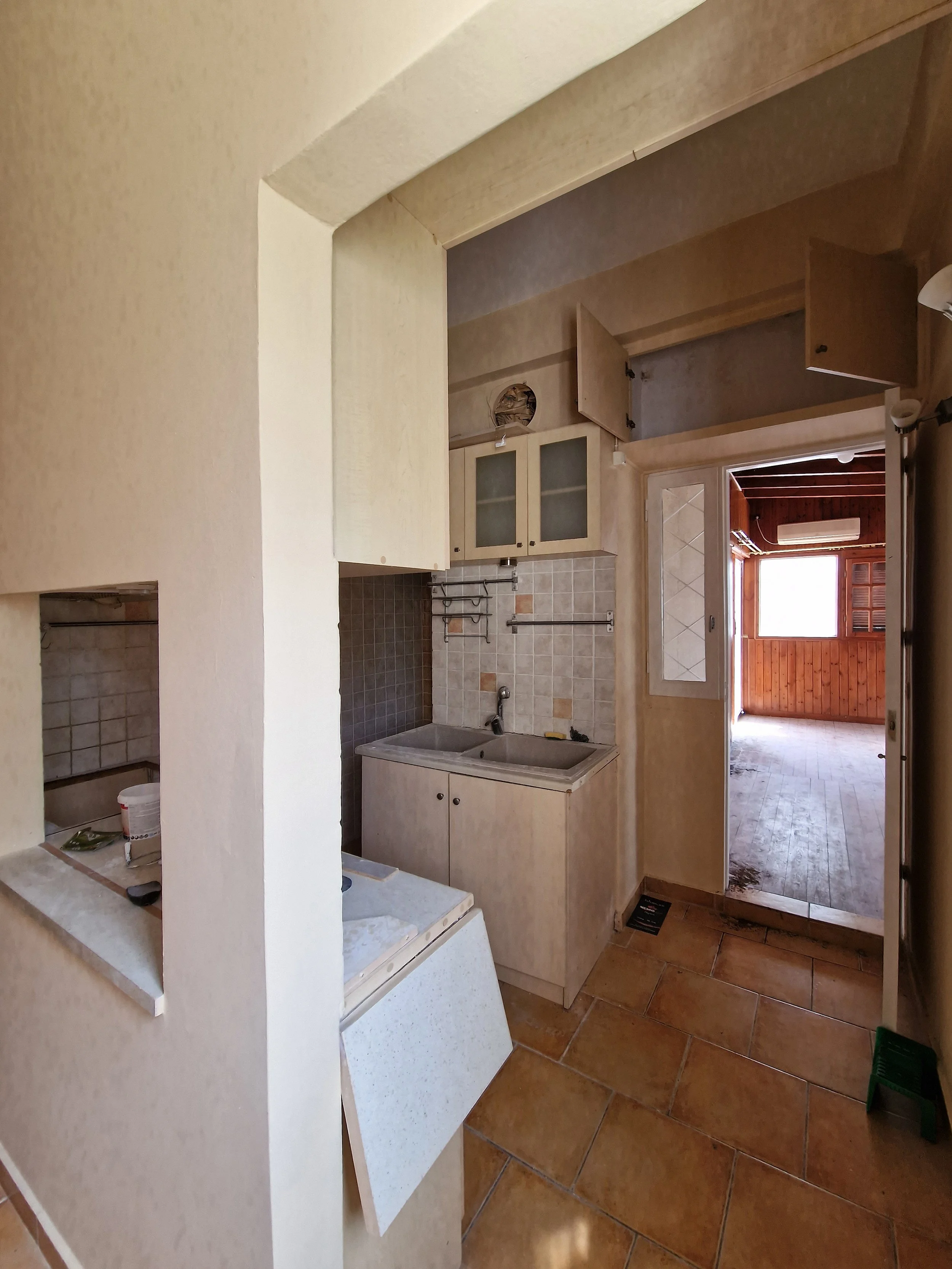 A view of an empty kitchen with beige walls, a small sink, upper cabinets with frosted glass doors, and an open doorway leading to a room with wooden paneling and an air conditioner.