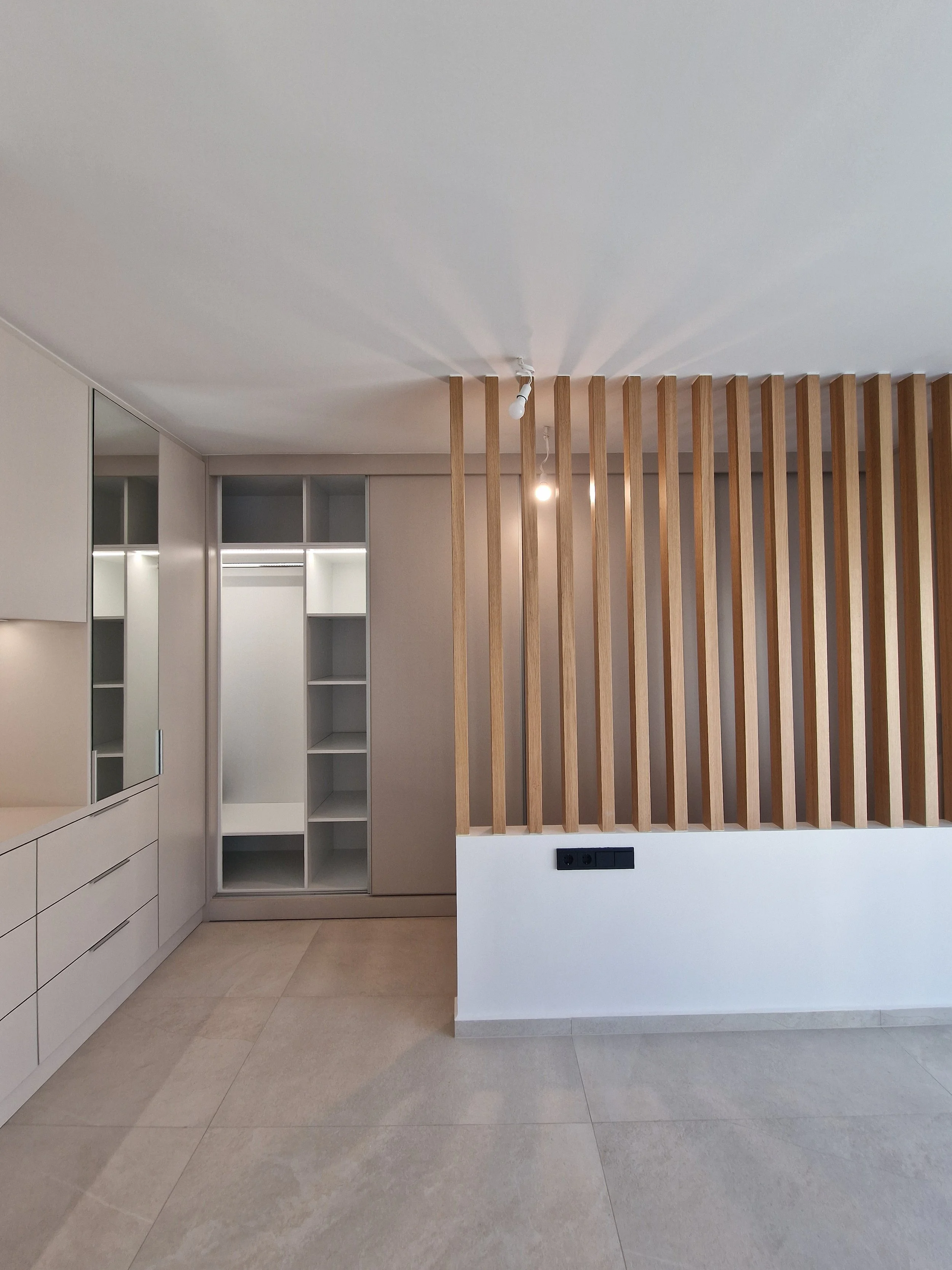 Interior of a modern room with a white cabinet with drawers on the left, a white shelving unit with open compartments, a wall with wooden slats dividing the space, and a beige tile floor.