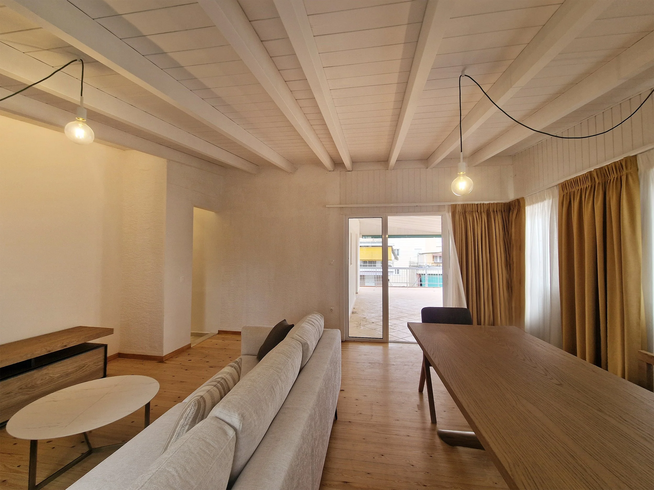 Living room with beige sofa, wooden coffee table, yellow curtains, and ceiling with exposed beams and hanging light bulbs.