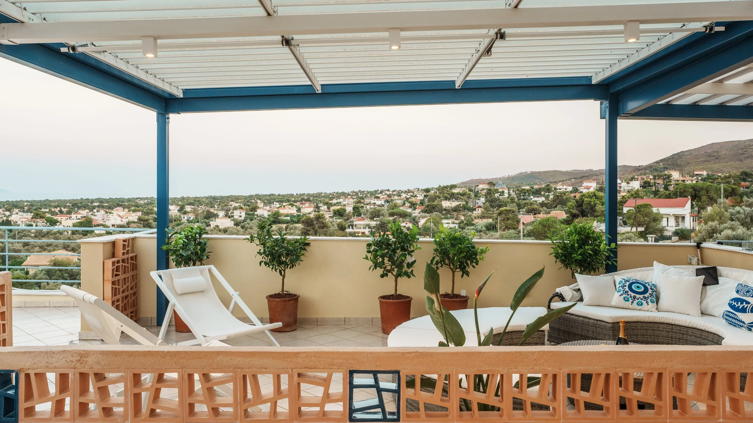Balcony with chairs and potted plants overlooking a cityscape with rolling hills in the background.