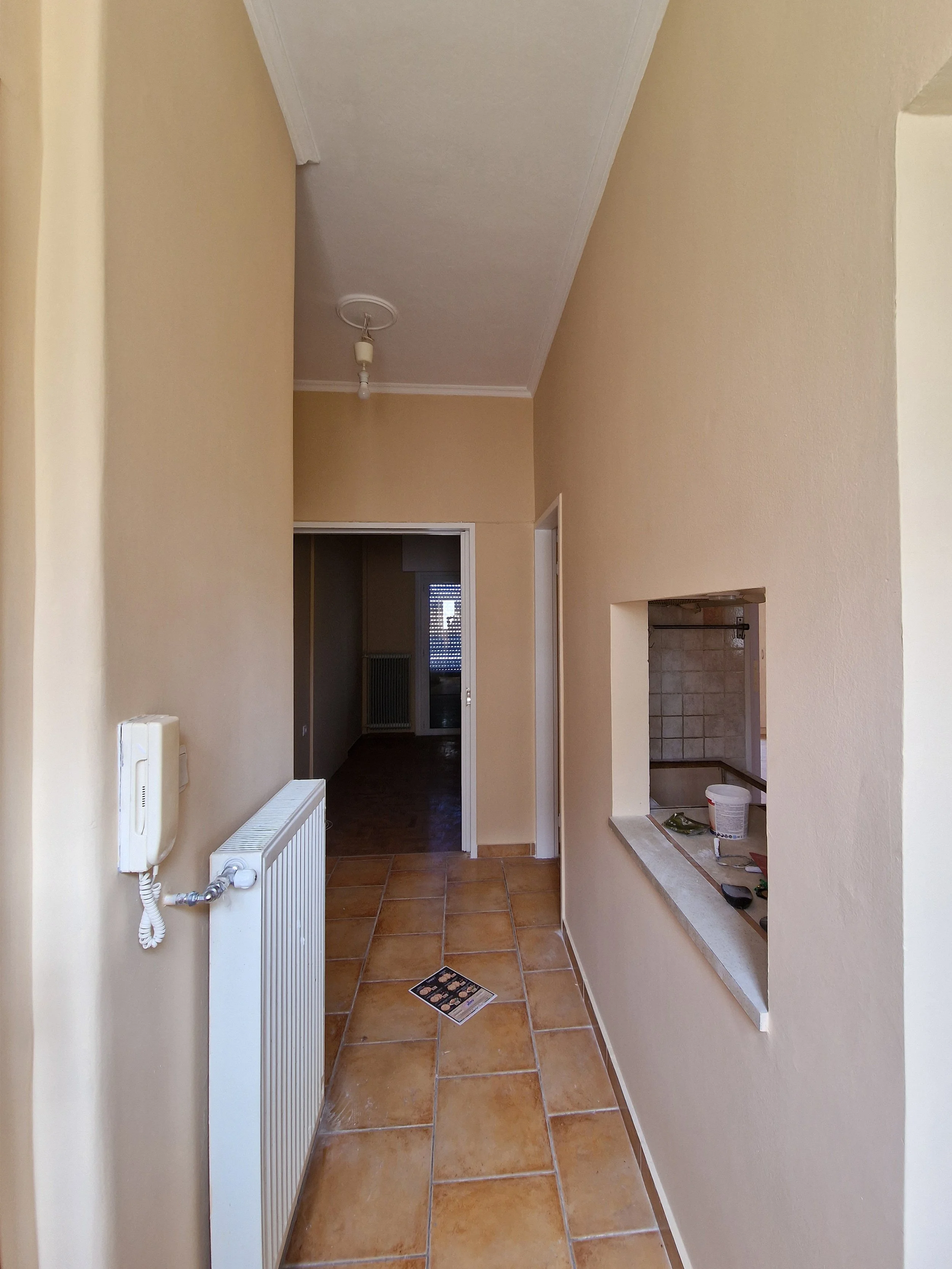 Empty hallway with orange-brown tile floor, beige walls, a radiator on the left wall, a doorway at the end, and a small opening in the right wall with construction tools and materials.