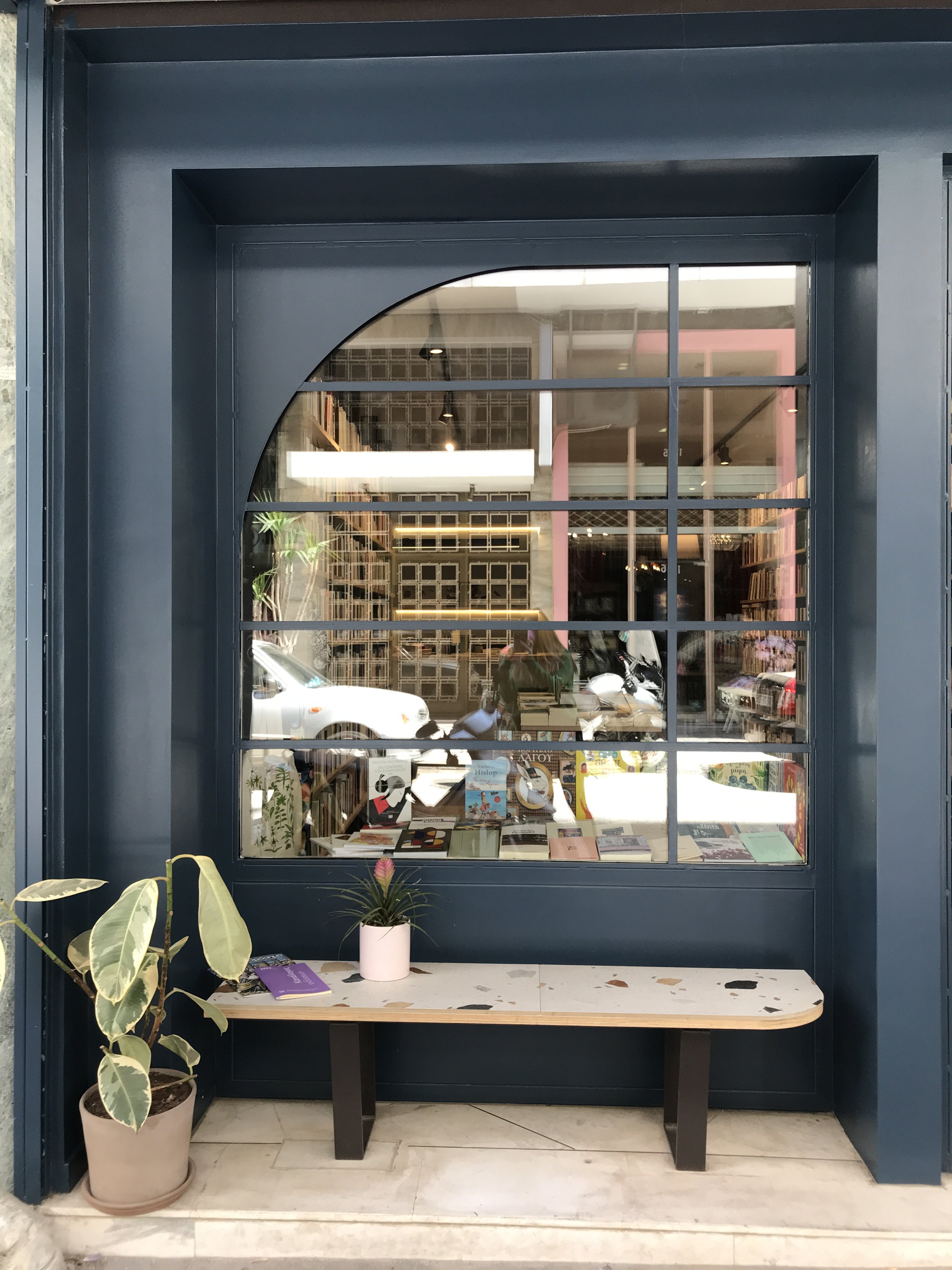 A bookstore window display with books inside, a pink potted plant on a table below the window, and a large potted plant with variegated leaves in front of the window. The window has a grid design, and the counter has a white top with black speckles.