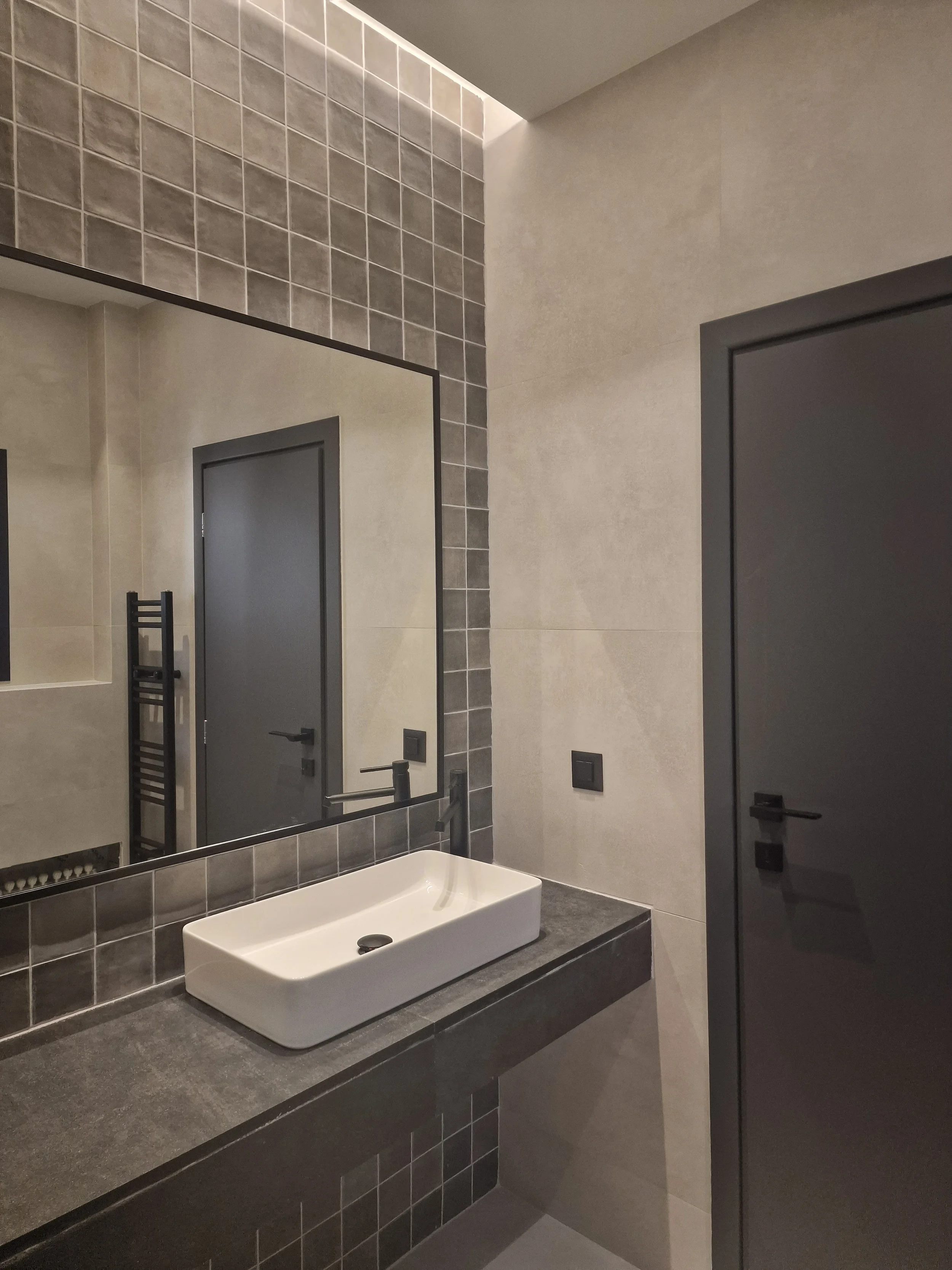 A modern bathroom with beige tiled walls, a black-framed mirror, a black door, a gray countertop, and a rectangular white sink.