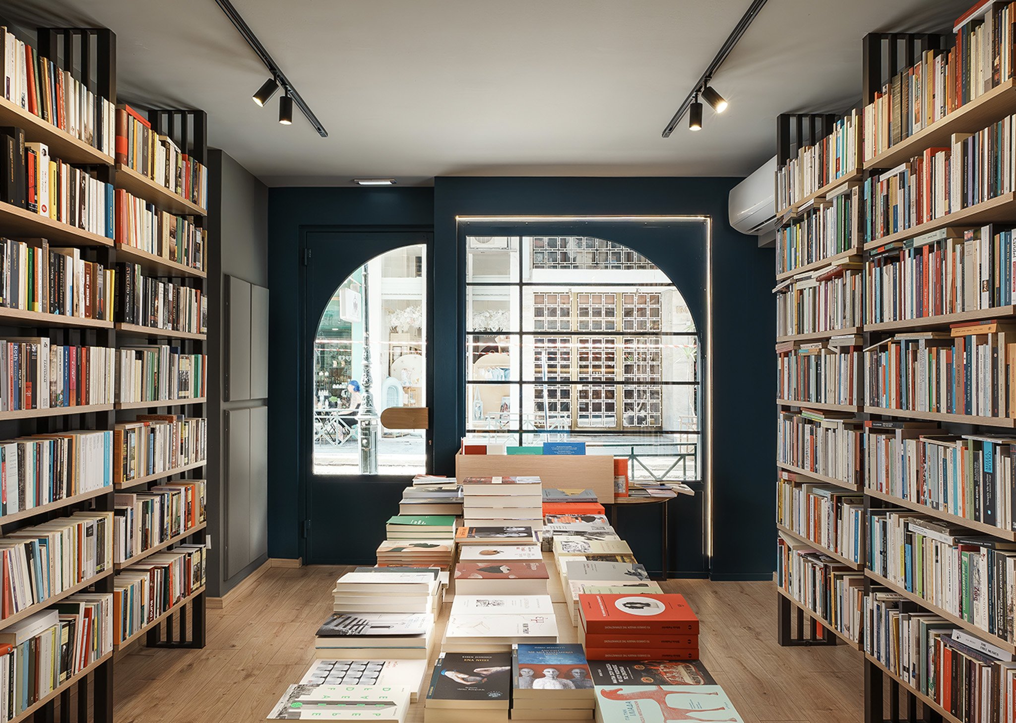 Interior of a bookstore with shelves filled with books, a table with books in the center, large windows allowing natural light, and ceiling track lighting.