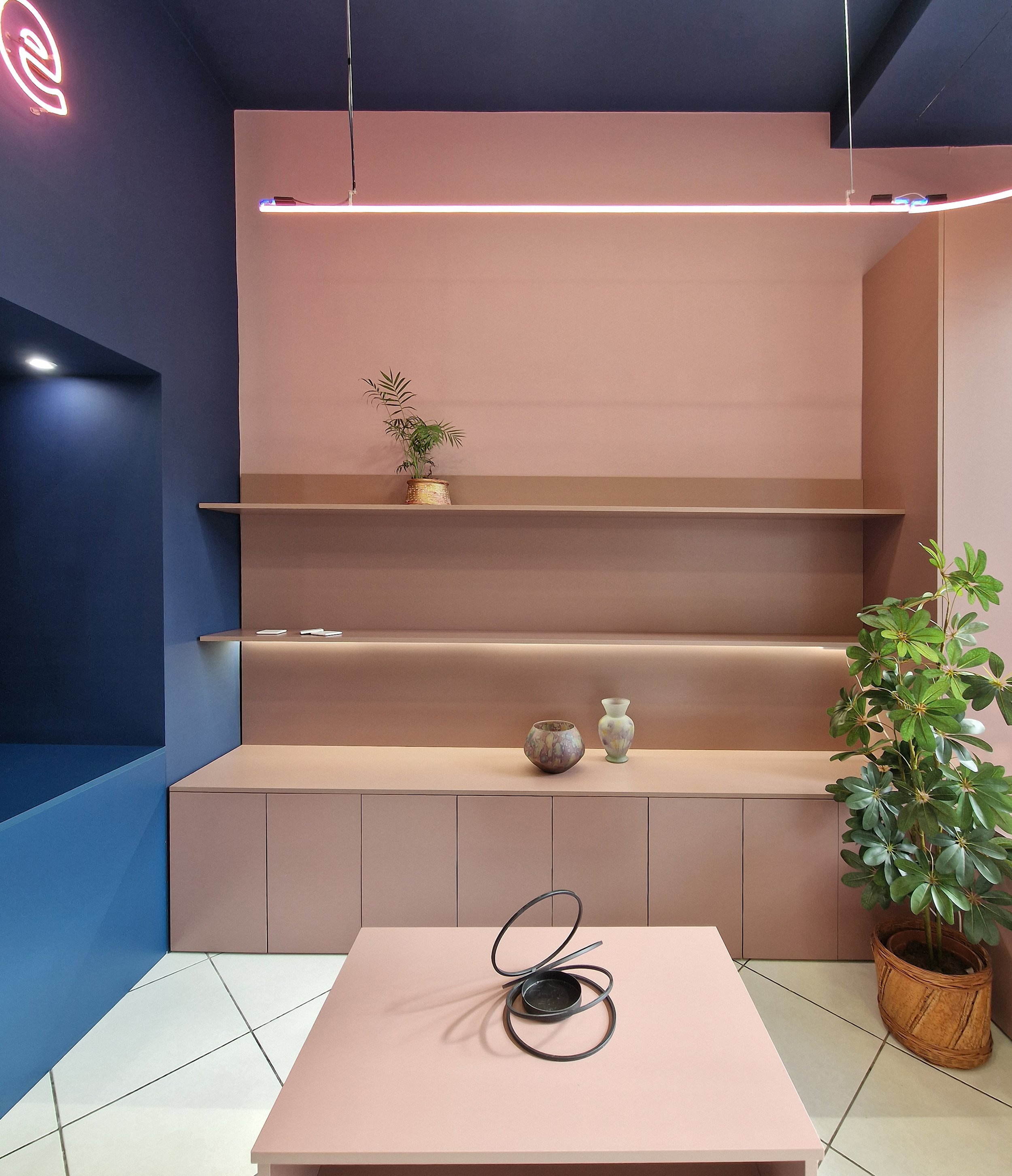 Interior scene with navy blue and pink walls, pink tables, a potted plant on a pink shelf, vases on the table, a headphone on the pink table, and a large leafy plant in a woven basket.