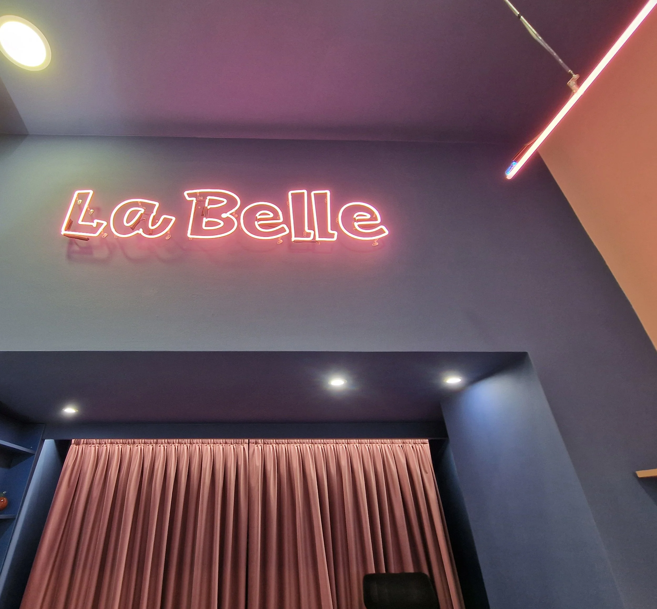 Interior of a venue with a neon sign that reads 'La Belle' and a pink curtain, with ceiling lights and a purple ceiling.
