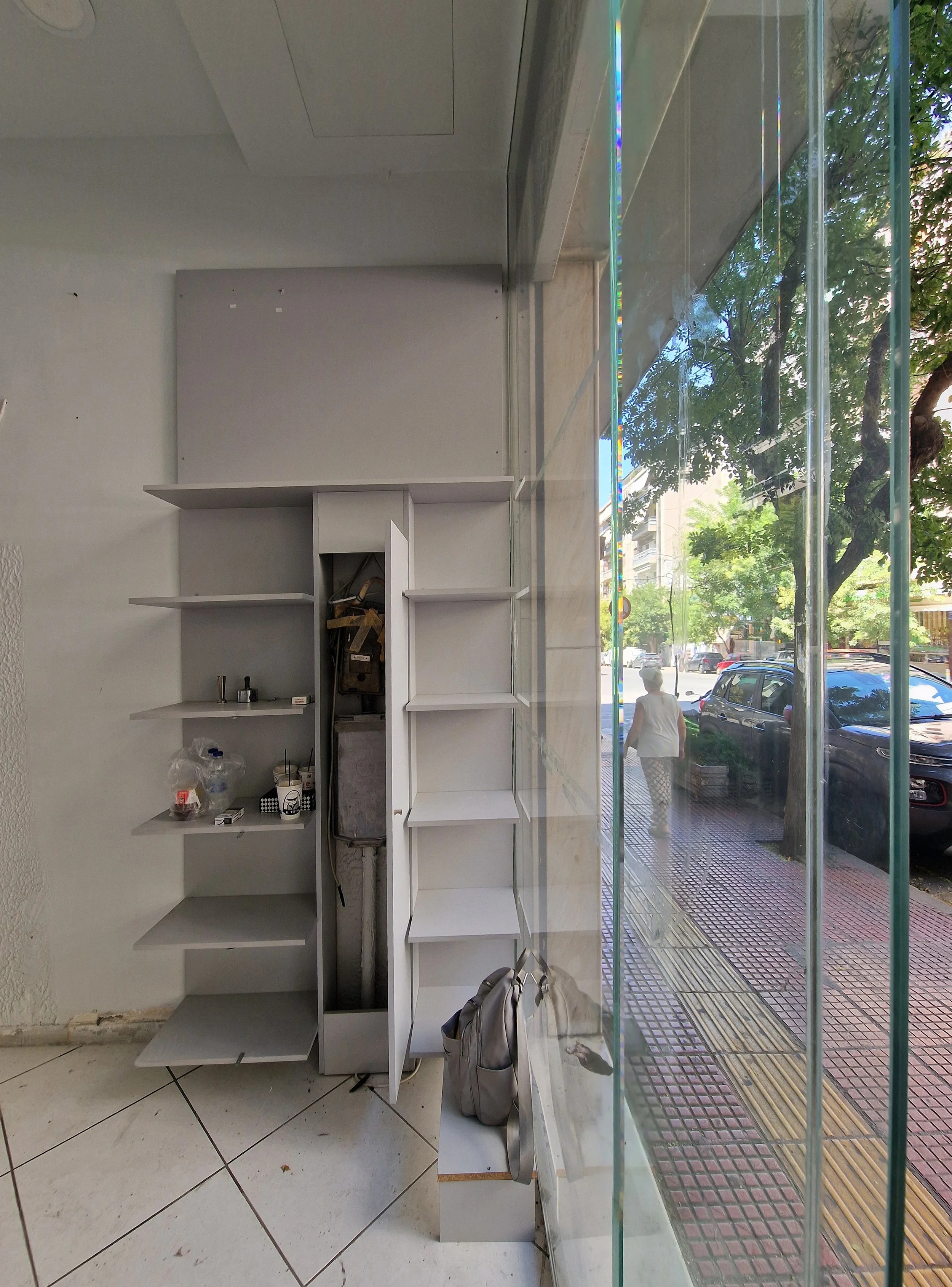 Empty retail space with white shelving units, a window with a view of a street with parked cars and trees, and a bag on the floor.