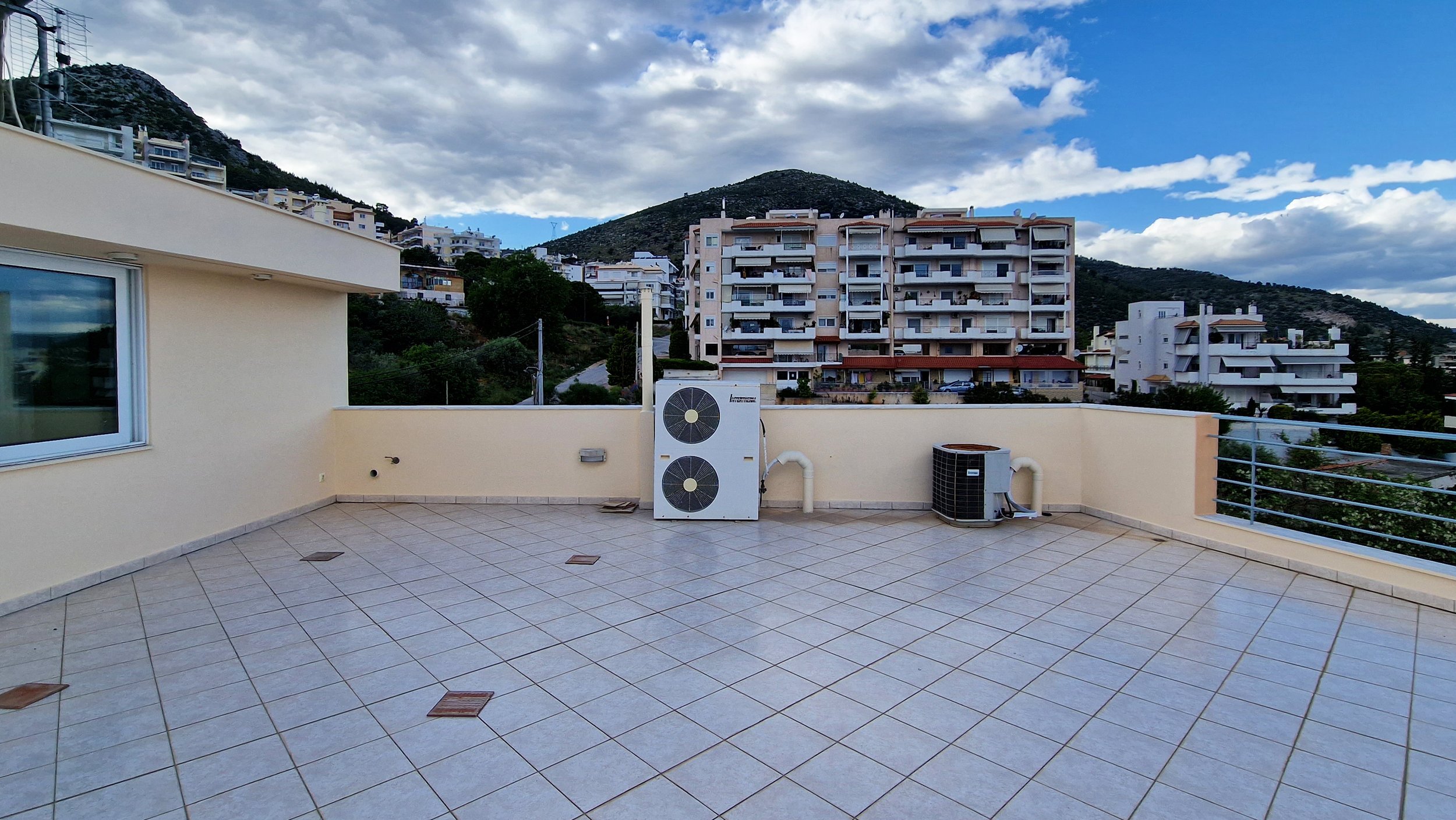 View from a balcony with beige tiled flooring and outdoor air conditioning units, overlooking multi-story residential buildings on hilly terrain under a partly cloudy sky.