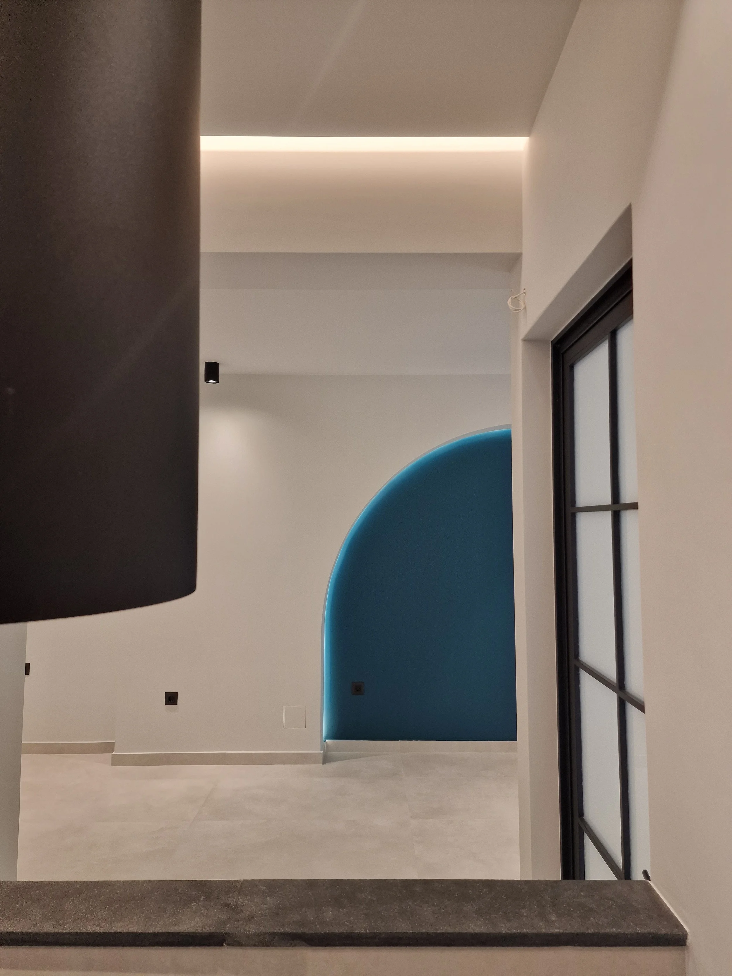 Modern interior space with a high ceiling, a blue curved wall feature, and a large window with black framing on the right. The room has light-colored flooring and smooth white walls with some electrical outlets visible.